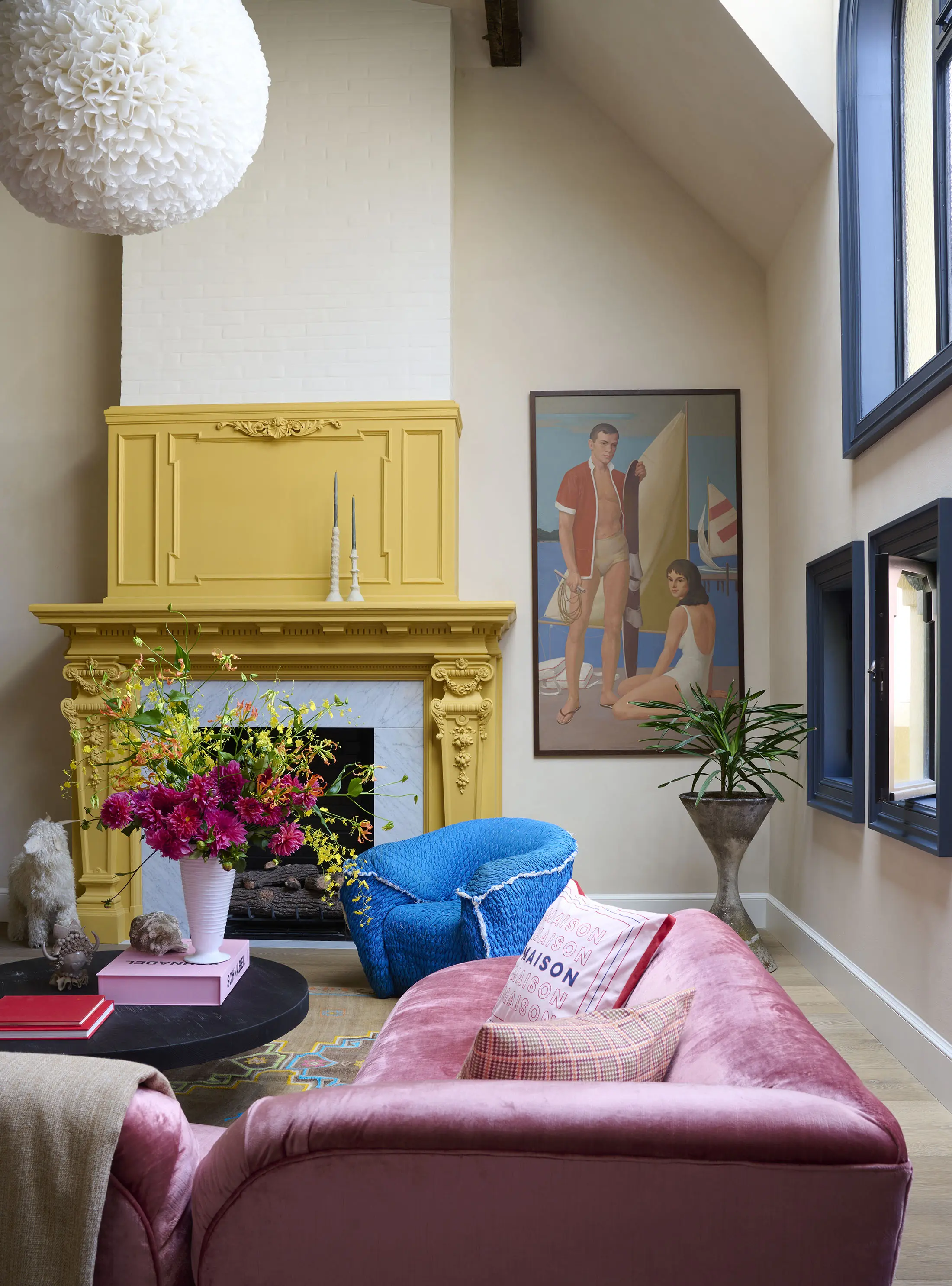 A stylish living room with a yellow ornate fireplace, a large painting of two people at the beach, and dark-framed windows. A pink velvet sofa with patterned pillows faces a blue armchair. A round coffee table holds a vibrant flower arrangement. A gray sculpture is nearby.