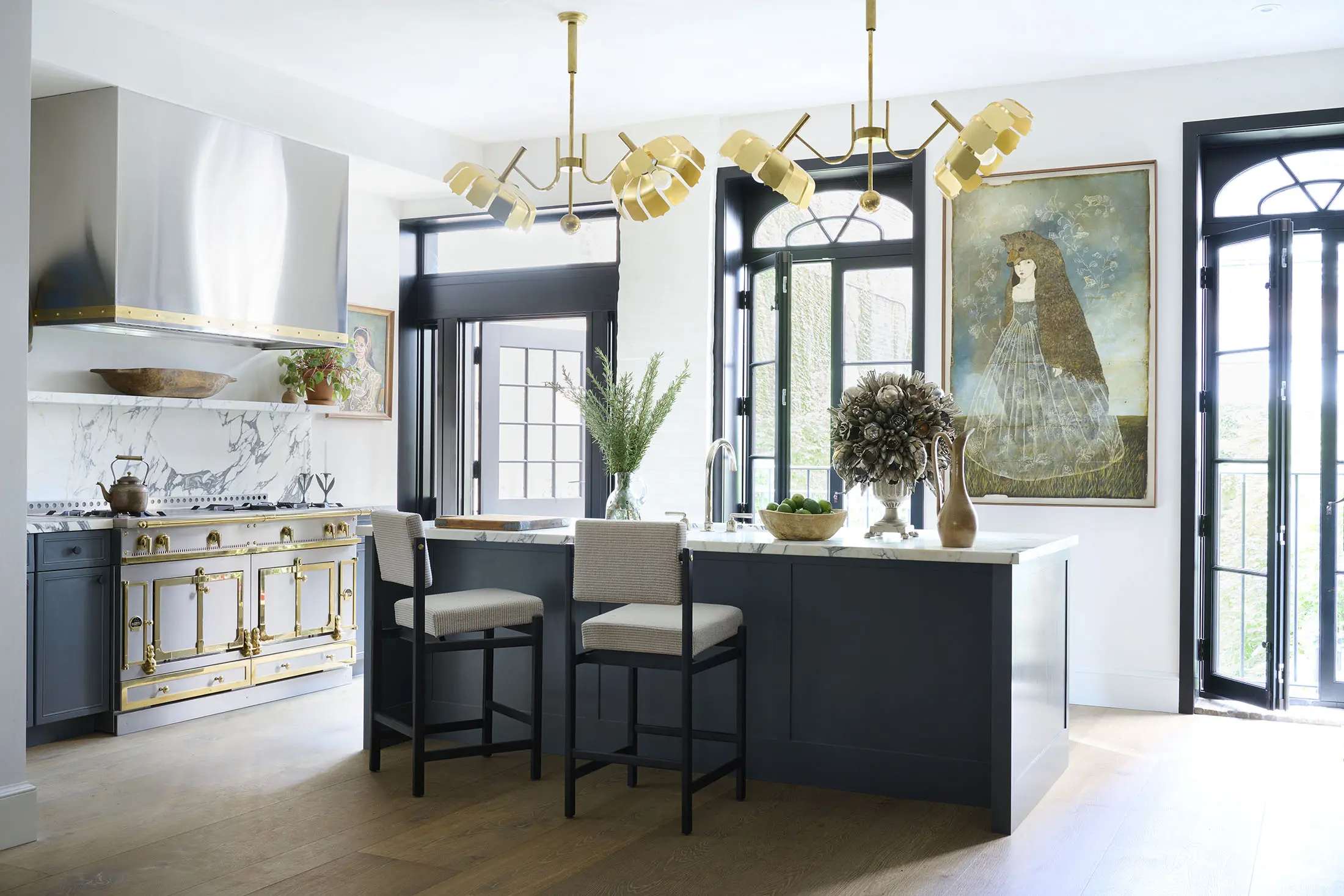 A modern kitchen with a navy island featuring two beige barstools. Above, two gold chandeliers hang. Black window and door frames complement a white wall. Artwork, vases with plants, and a bowl of apples decorate the space. The floor is light wood.