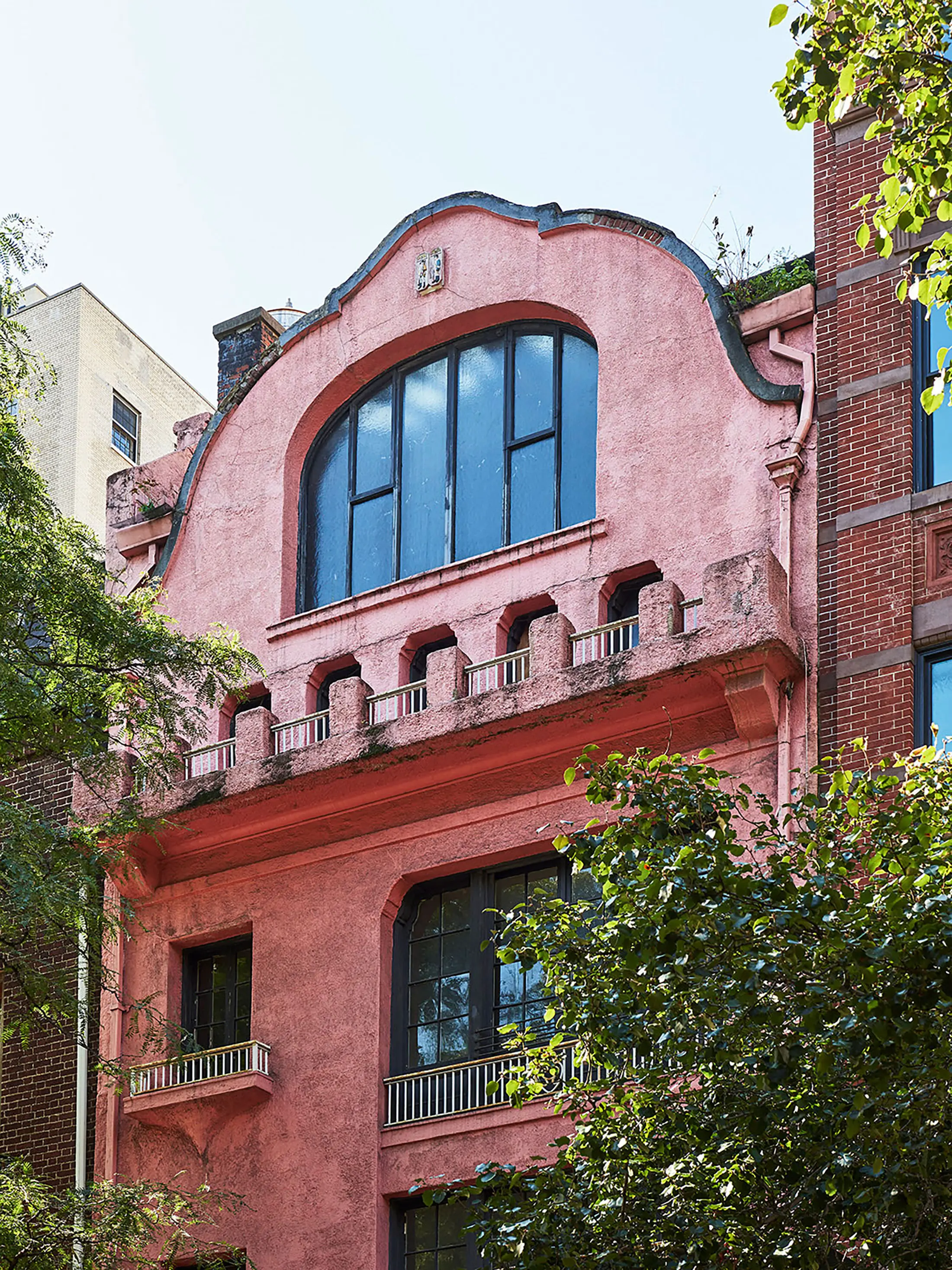 A modern kitchen with a navy island featuring two beige barstools. Above, two gold chandeliers hang. Black window and door frames complement a white wall. Artwork, vases with plants, and a bowl of apples decorate the space. The floor is light wood.