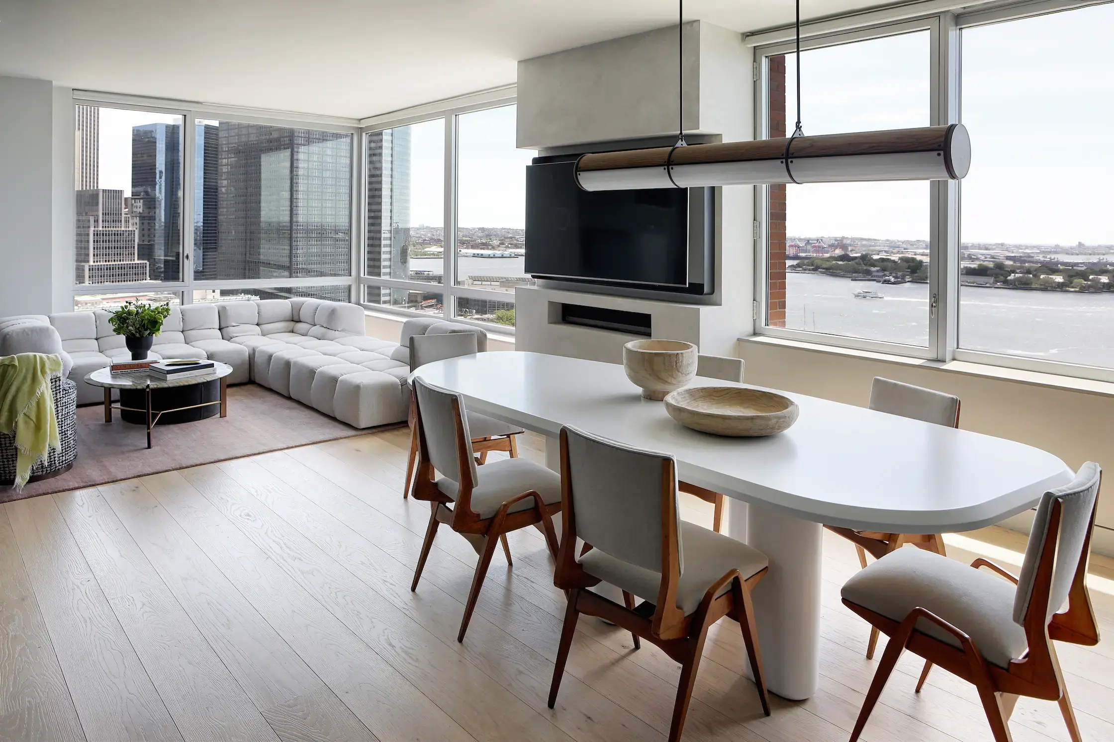 A modern, open-concept living and dining area in a high-rise apartment with floor-to-ceiling windows offering a view of the city skyline and waterfront. The space features a white oval dining table surrounded by six upholstered wooden chairs, a contemporary sectional sofa in a neutral tone, a round coffee table, and a built-in television mounted on a sleek accent wall. Light wood flooring and minimalist decor enhance the bright, airy ambiance.