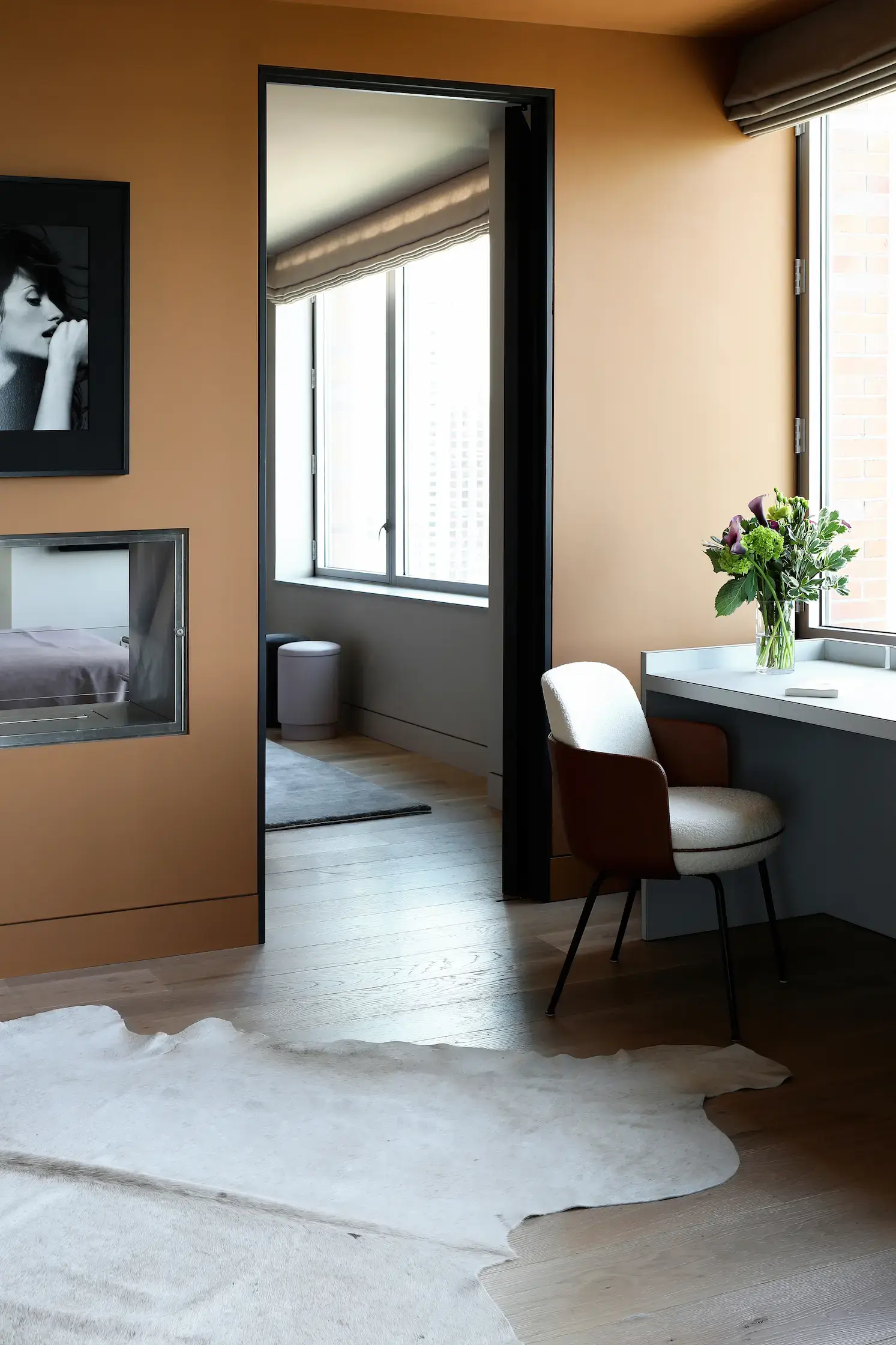 A stylish bedroom corner featuring a built-in desk by a large window with natural light streaming in. A modern upholstered chair with a curved back and metal legs sits at the desk, which holds a vase of fresh flowers. The room has warm-toned walls, light wood flooring, and a white cowhide rug. A black-framed portrait hangs on the wall, and a sleek, glass-enclosed fireplace offers a glimpse into an adjoining bedroom with a neatly made bed. A doorway leads to a bright sitting area with a soft rug and a small ottoman.