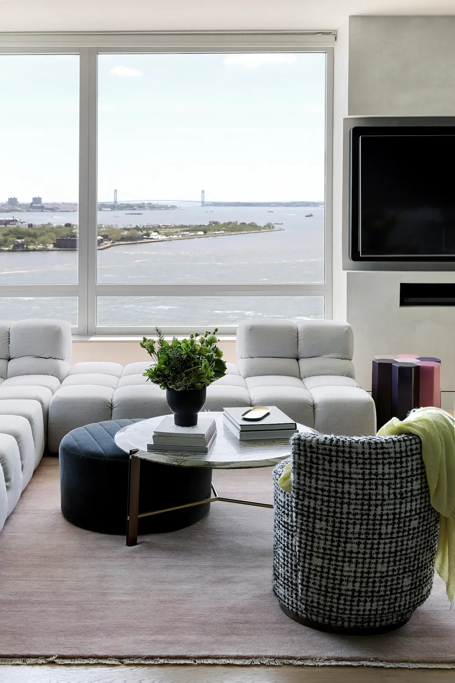 A modern living space with a panoramic waterfront view through a large window. The room features a plush, tufted white sectional sofa, a round marble coffee table with a gold metal base, and a black upholstered ottoman. A black and white tweed accent chair with a yellow throw blanket is positioned nearby. The coffee table is adorned with a potted green plant and stacked books. A mounted television is on a sleek, built-in wall unit. A muted pink area rug covers the light wood flooring, creating a cozy yet sophisticated ambiance. In the background, a bridge and greenery are visible across the water.