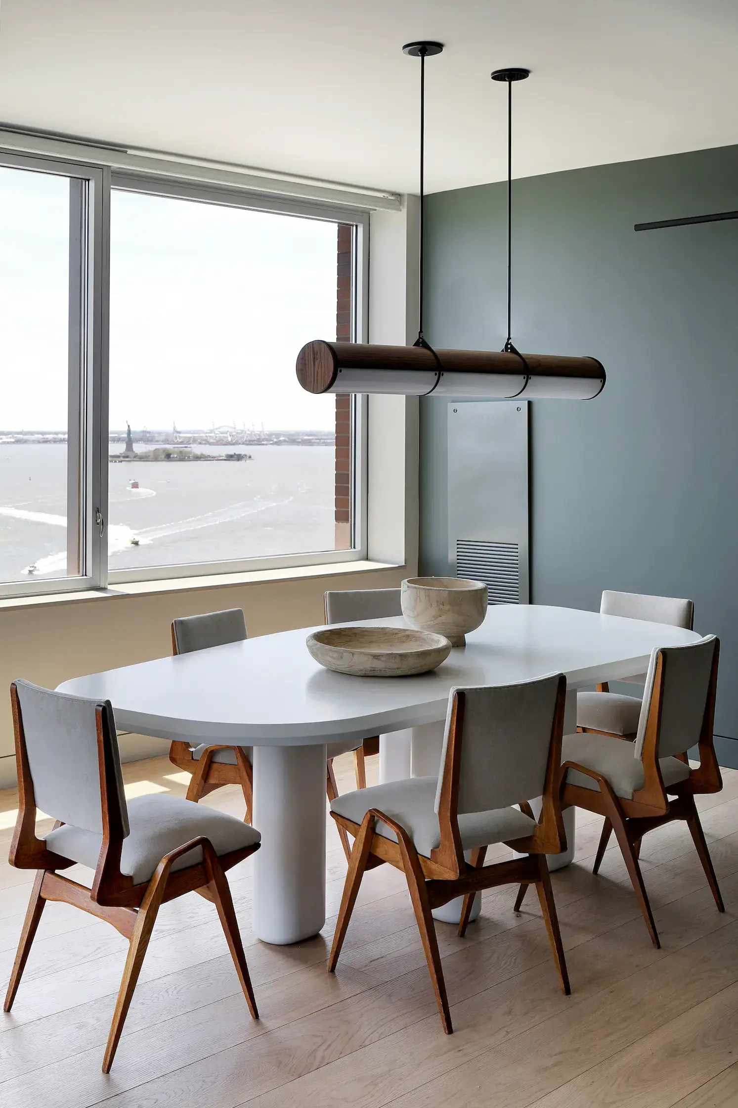Modern dining room with a large white oval table, surrounded by six wooden chairs with light gray cushions. A minimalist cylindrical pendant light hangs above. Large window offers a view of boats and a cityscape across the water. Light wood flooring.