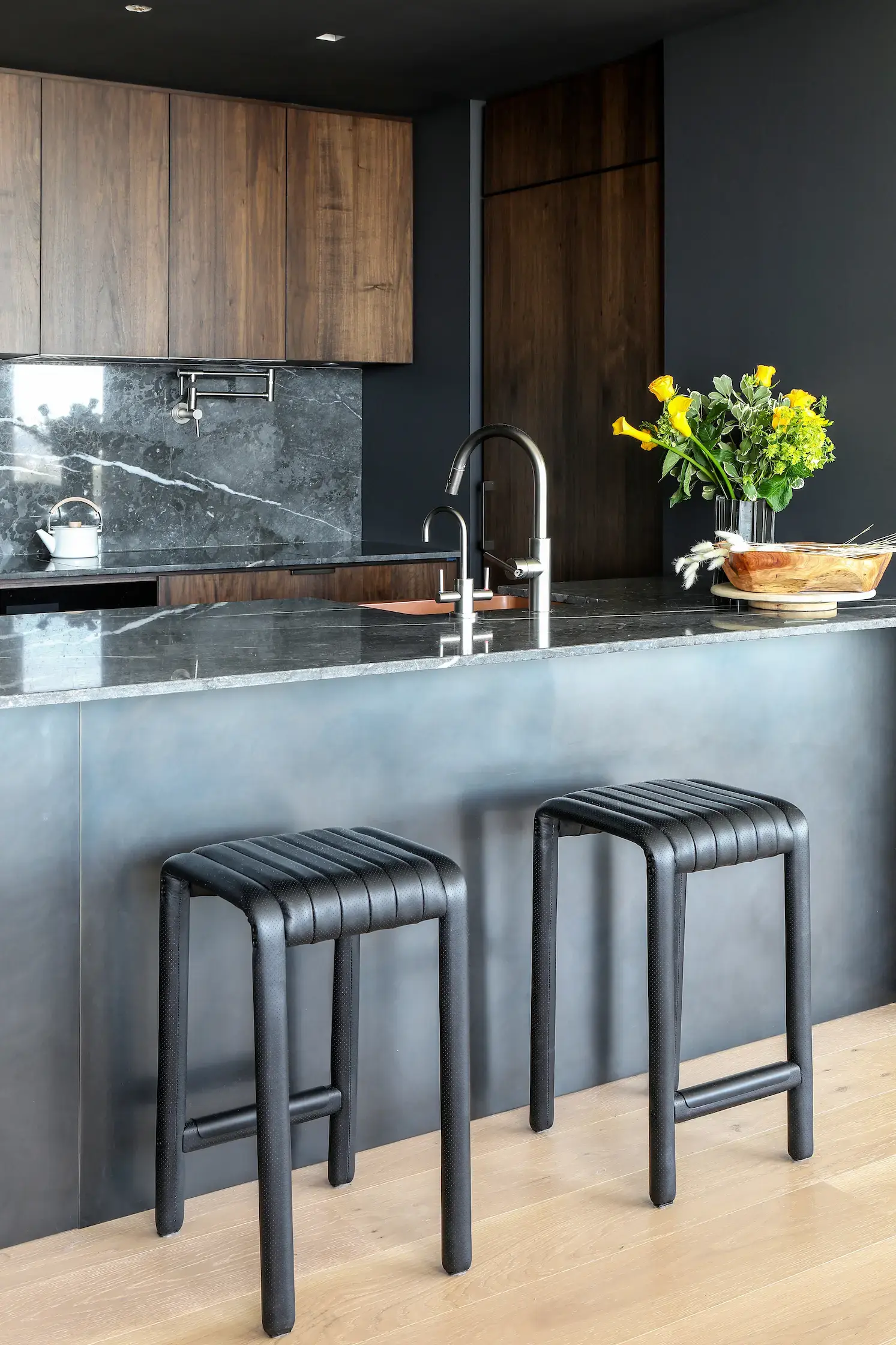 Modern kitchen with dark wood cabinets, black marble backsplash, and countertops. A silver faucet is mounted on the island, with two black bar stools in front. A vase with yellow flowers and a wooden bowl decorate the counter. Light wooden flooring contrasts the dark decor.