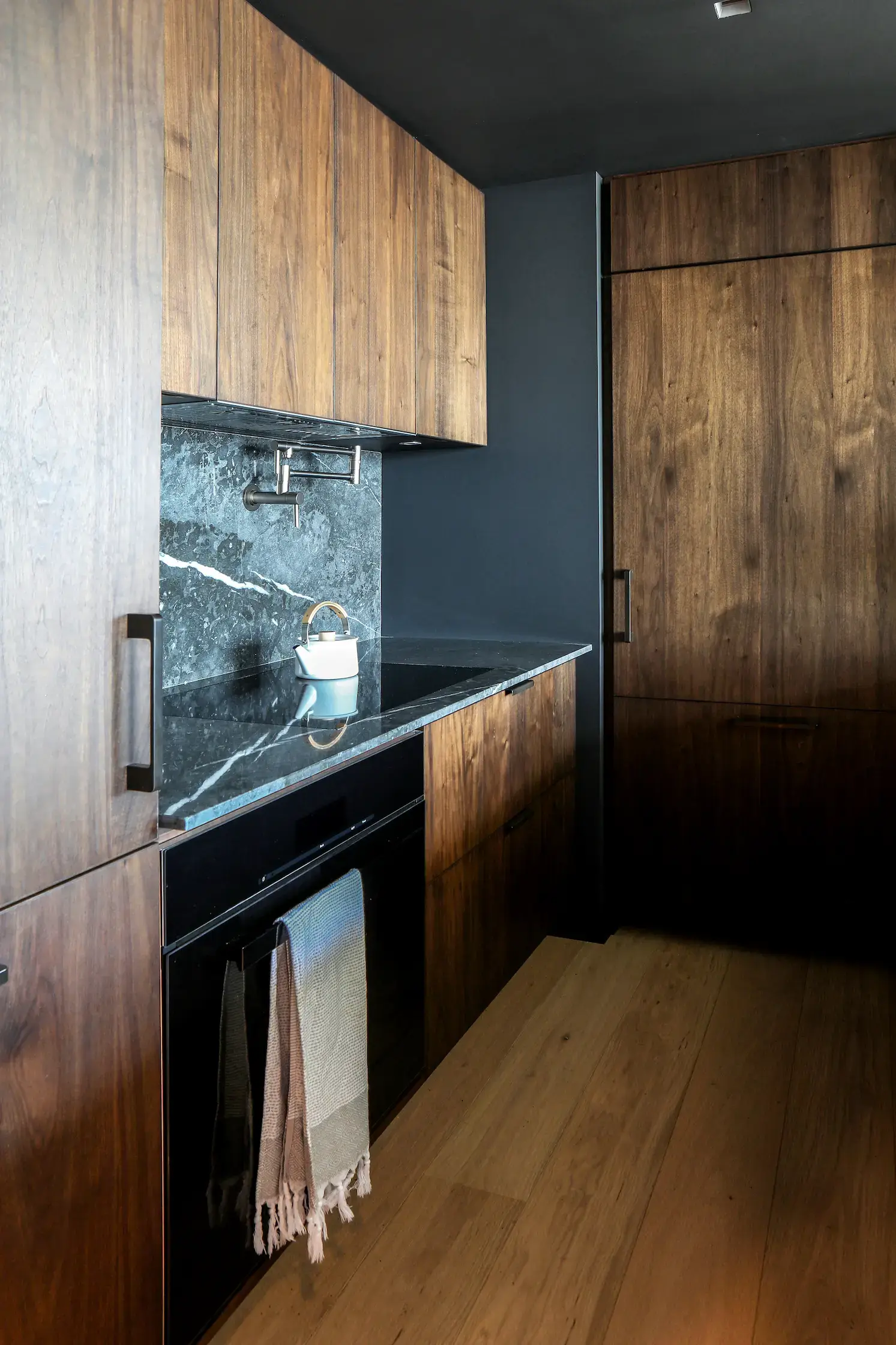 Modern kitchen with dark wood cabinets and black marble backsplash. Built-in oven with a hanging dish towel in front. A pot rests on the marble countertop under a wall-mounted faucet. Light wood flooring contrasts with the dark cabinetry and walls.