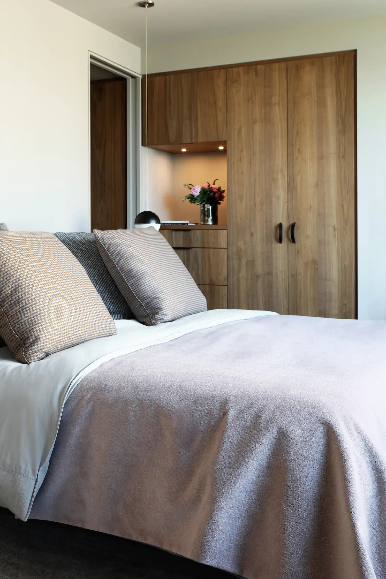 Modern bedroom with a neatly made bed featuring gray and beige pillows and a light brown blanket. A wooden wardrobe and a small shelf with a vase of flowers are in the background. Soft lighting highlights the room’s minimalist design and warm, natural tones.