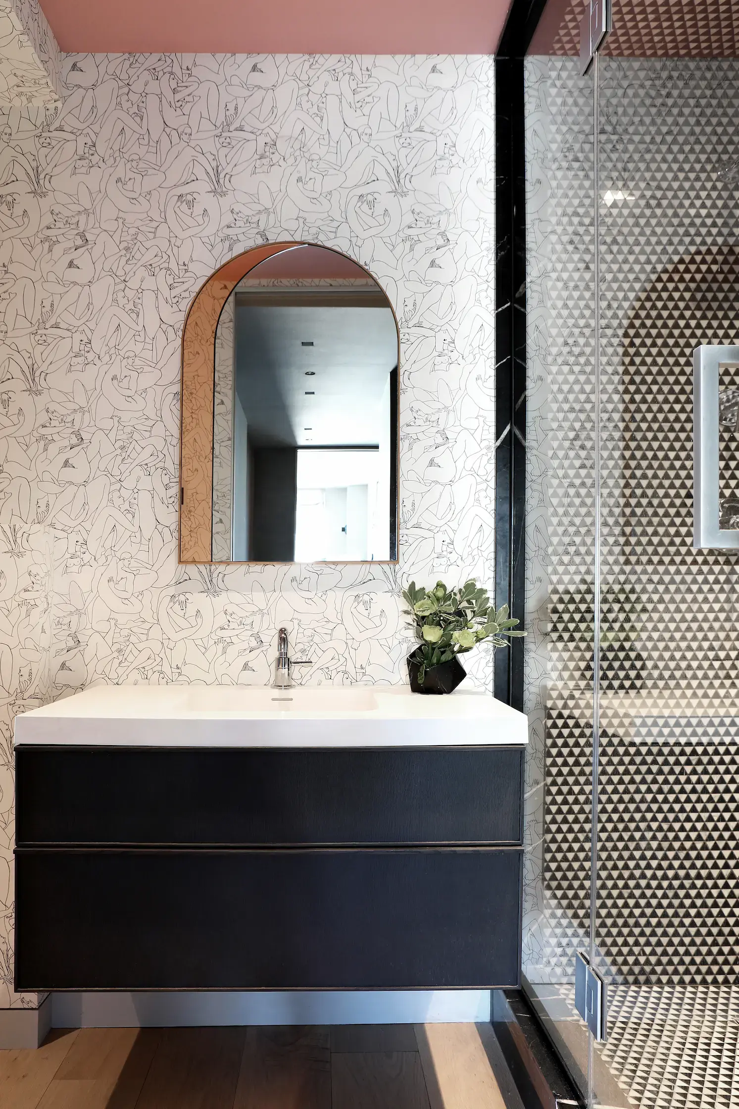 Modern bathroom with a floral-patterned wall and an arched mirror above a dark, floating vanity. A potted plant sits on the sink counter. To the right, theres a glass shower enclosure with a geometric design. The wooden floor adds warmth to the space.