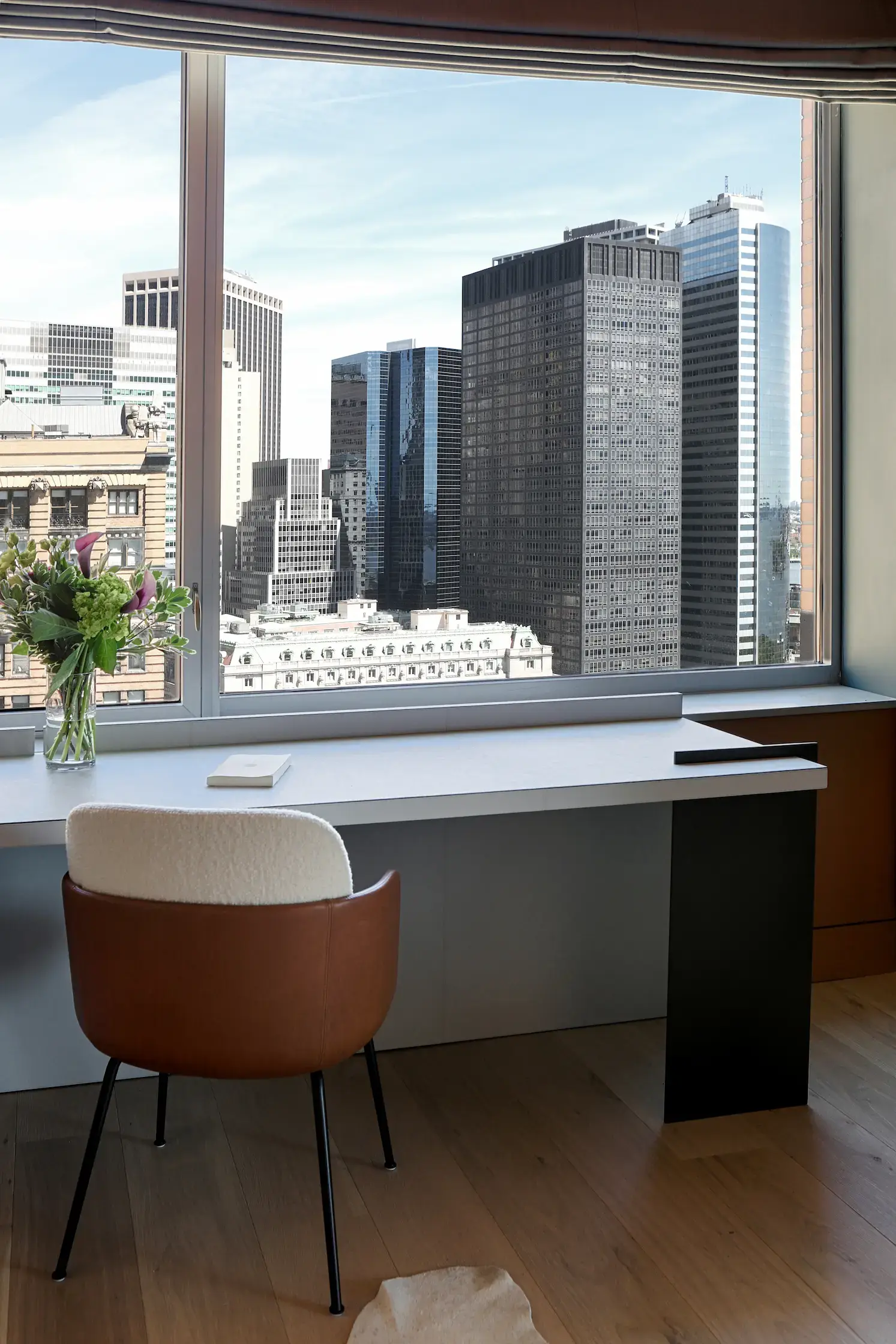 A modern office space with a desk facing a large window showing a cityscape of tall buildings under a clear sky. On the desk are a closed laptop, a small notepad, and a vase with colorful flowers. A brown chair with a white cushion is neatly positioned at the desk.