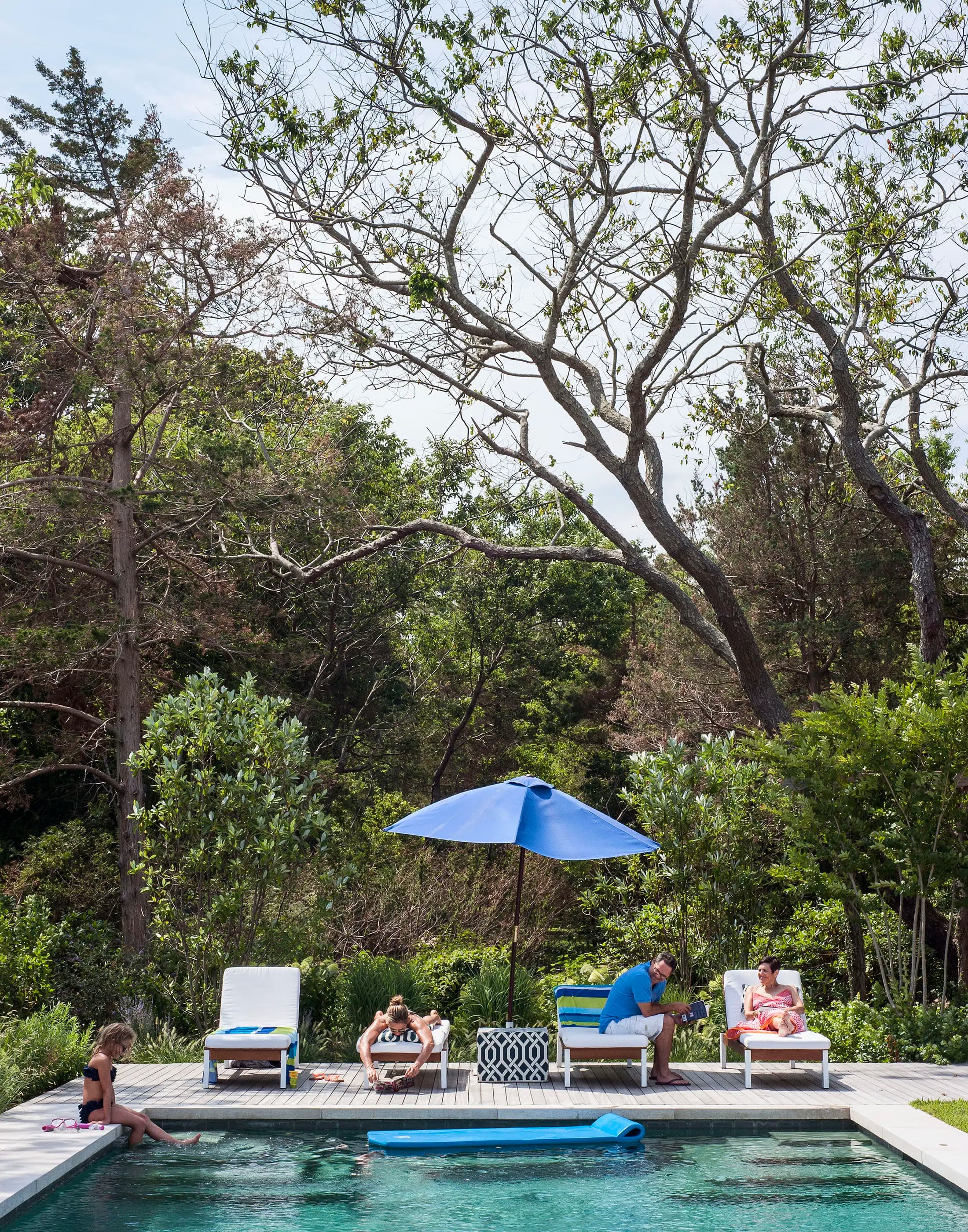 People relaxing by a pool surrounded by dense trees. One person sits in a chair under a large blue umbrella, while others are on sun loungers. A blue float is in the pool. Bright summer day with clear skies, and lush greenery frames the scene.