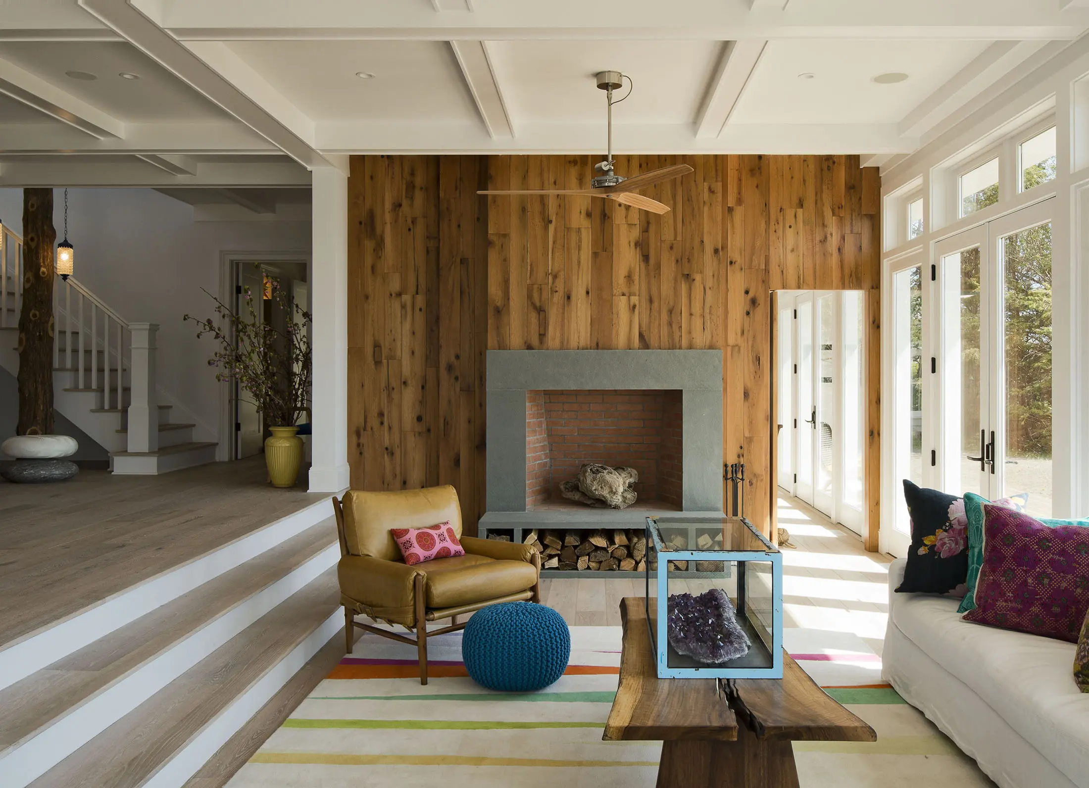 A cozy living room features a wooden accent wall with a gray fireplace, adorned with a red brick interior. There are chairs with colorful cushions, a blue pouf, and a multicolored striped rug. Large windows on the right let in ample light, illuminating the space.