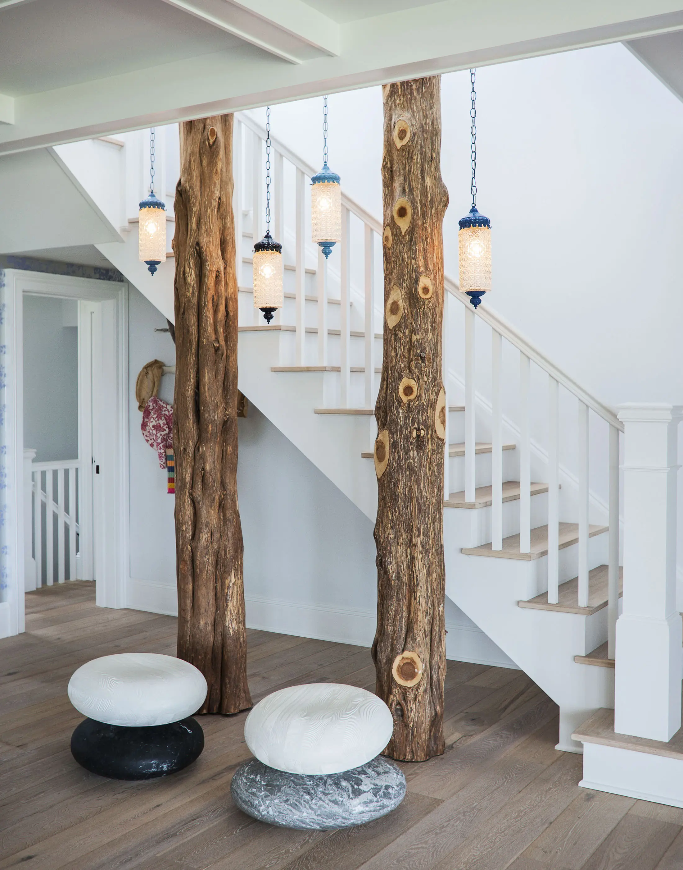 Interior view of a staircase with rustic wooden pillars resembling tree trunks. Hanging lanterns with frosted glass illuminate the space. Two stone-like round stools, one dark gray and one white, are on the wooden floor. The walls are light-colored, creating a natural aesthetic.