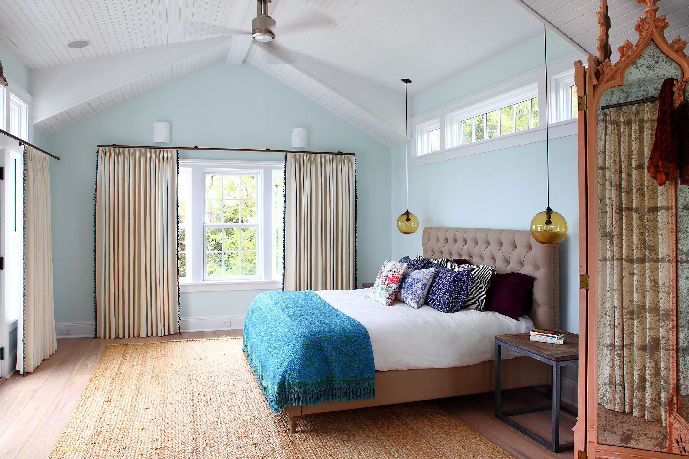 A bedroom with light blue walls, featuring a beige tufted bed with white, purple, and blue pillows. A teal blanket is draped over the bed. Two gold pendant lights hang above. Large windows with cream curtains are on the far wall. The room has a high ceiling fan and a wooden floor with a rug.