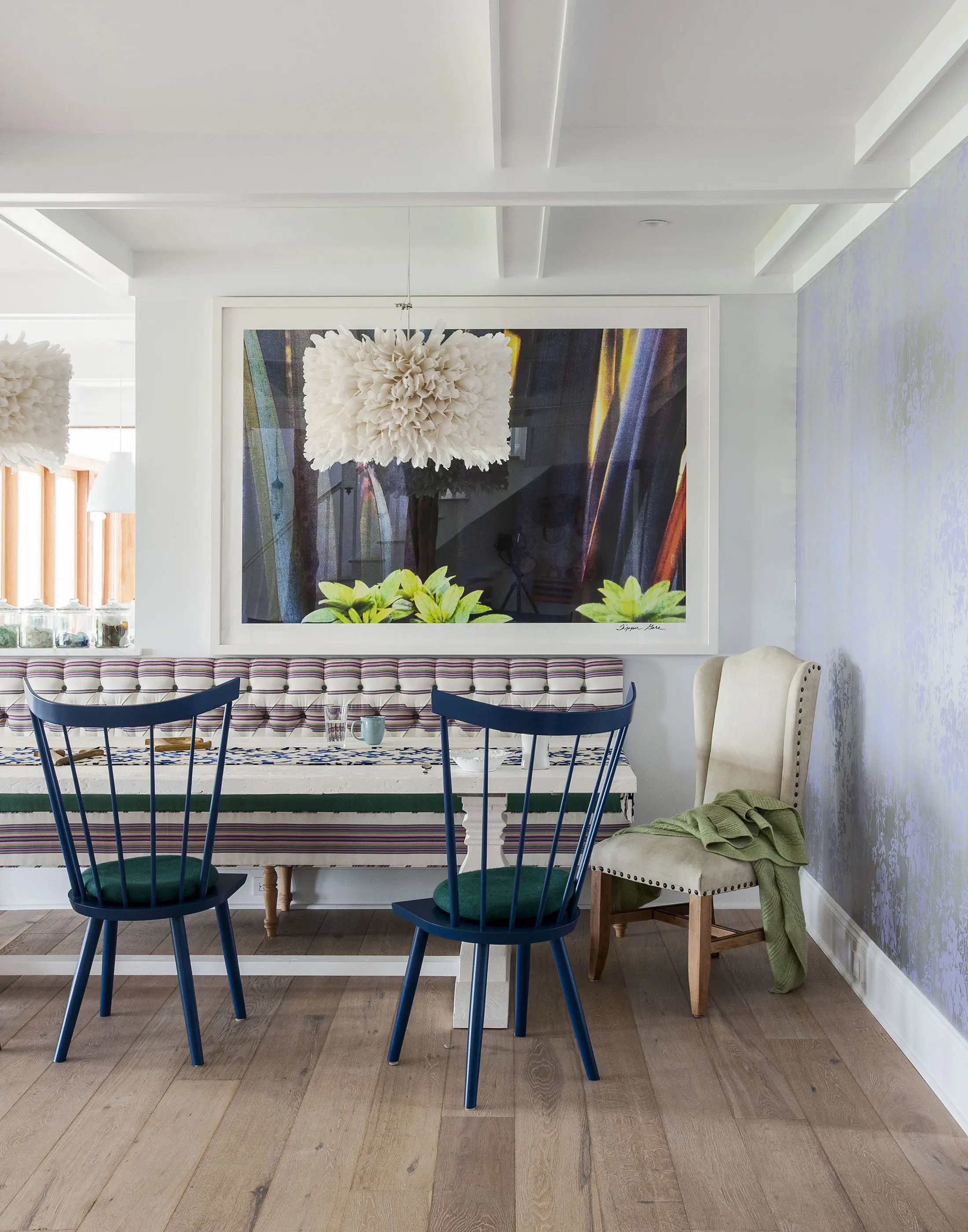 A modern dining area features a long, patterned bench with round cushions, two dark blue chairs with green seats, and a beige upholstered chair with a green fabric draped over. Above, a large abstract artwork and a white feathered pendant light hang on the wall amidst light purple wallpaper.