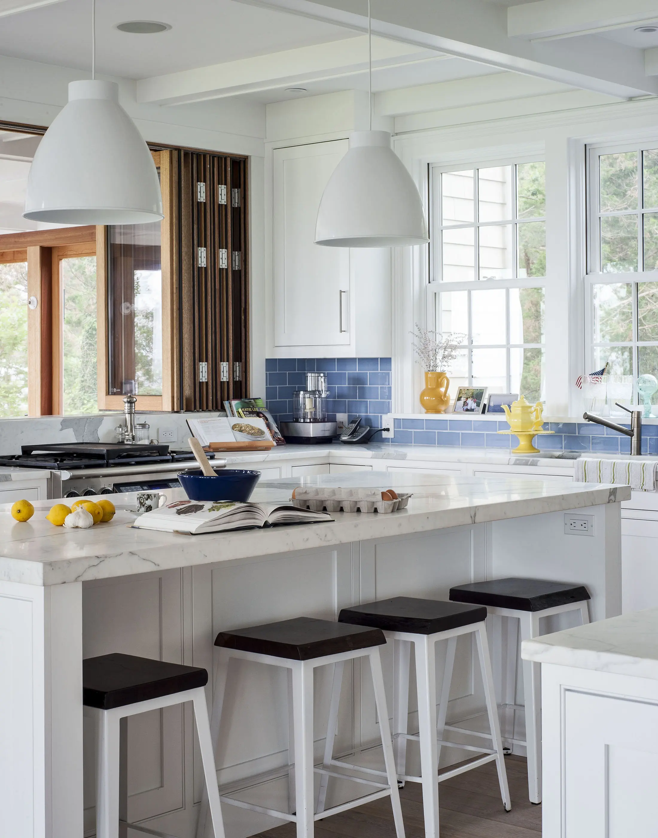 Modern kitchen with white cabinets and marble countertops. A blue tile backsplash contrasts with white walls. Three black-topped stools sit at the central island, which has lemons, a cookbook, and eggs. Two white pendant lights hang above. Sunlight streams through large windows.