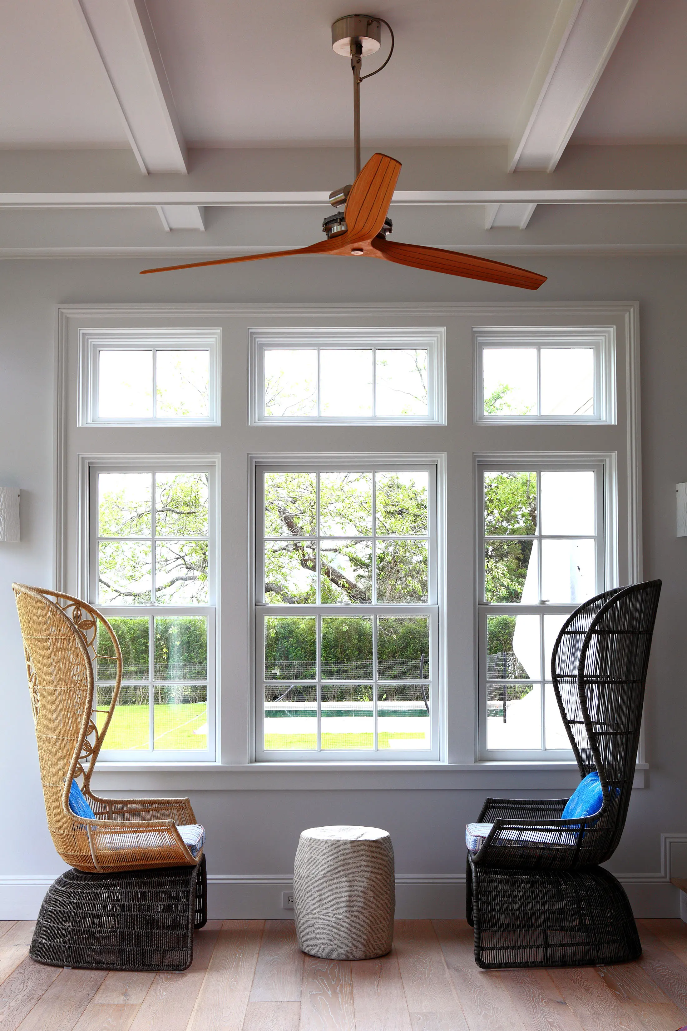 Two tall wicker chairs face each other, one tan and the other black, flanking a small round stone table. They are beside large windows showing a green lawn and trees. Above, a wooden ceiling fan contrasts with the white beamed ceiling. The floor is light wood.