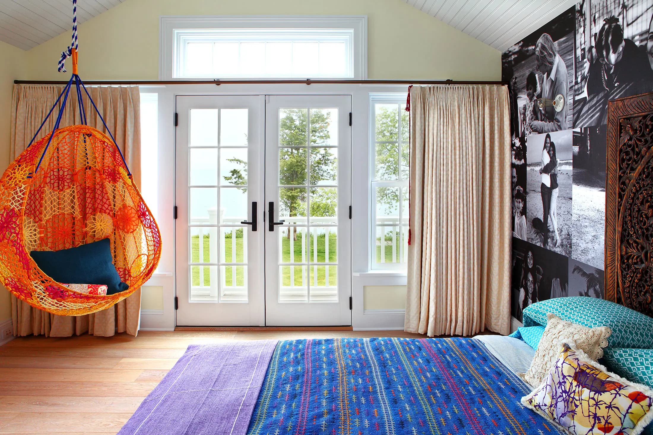 A colorful, airy bedroom features a crochet hanging chair with a cushion, wooden floor, and a vibrant bedspread. A wall displays black-and-white photos. Large curtained doors open to a scenic view of trees and water, with soft natural light filling the room.