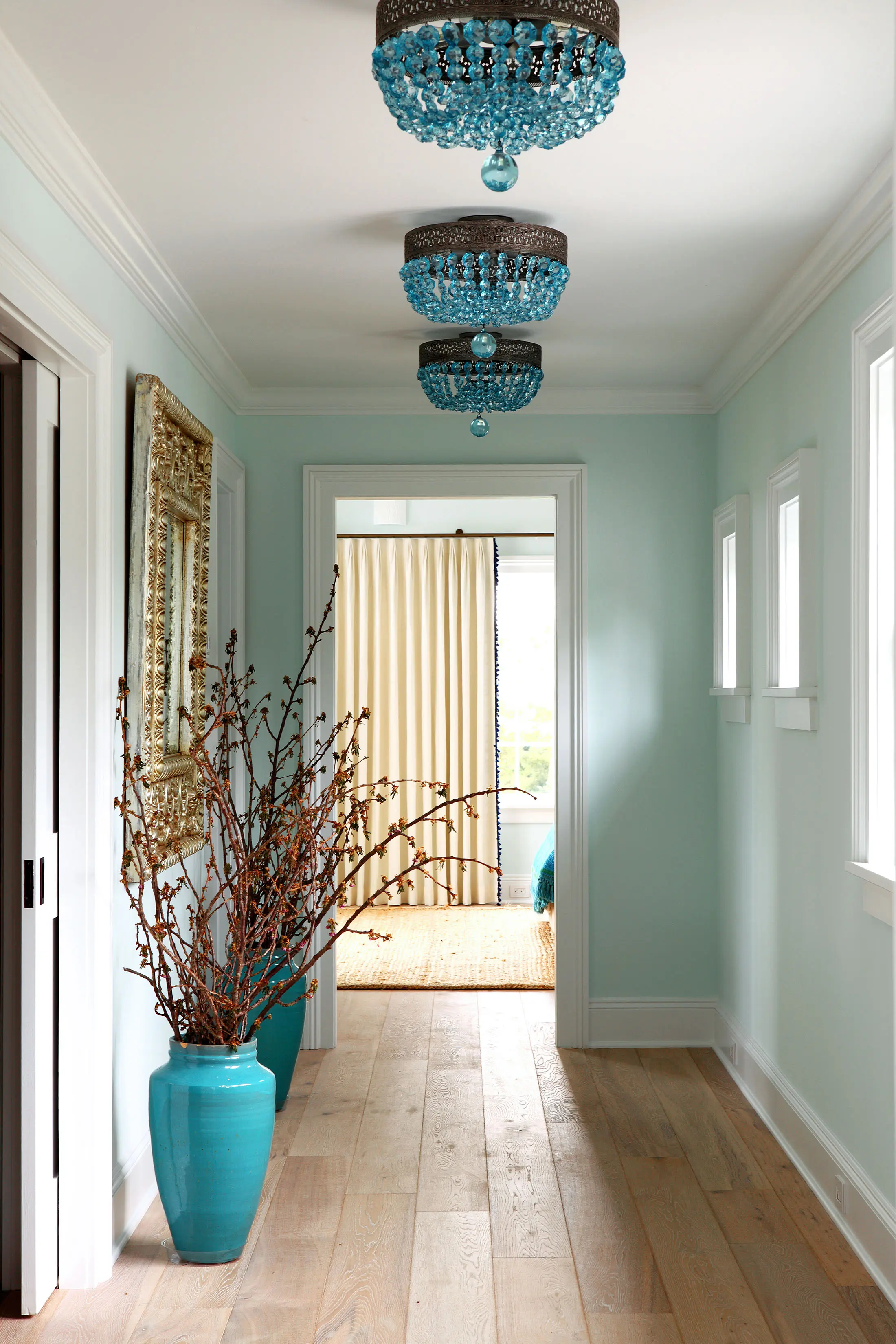 A hallway with light blue walls and three turquoise chandeliers. The floor is light wood, and large turquoise vases with branches sit on the left. A gold-framed mirror is above the vases. At the end of the hallway, cream-colored curtains and a woven mat are visible.