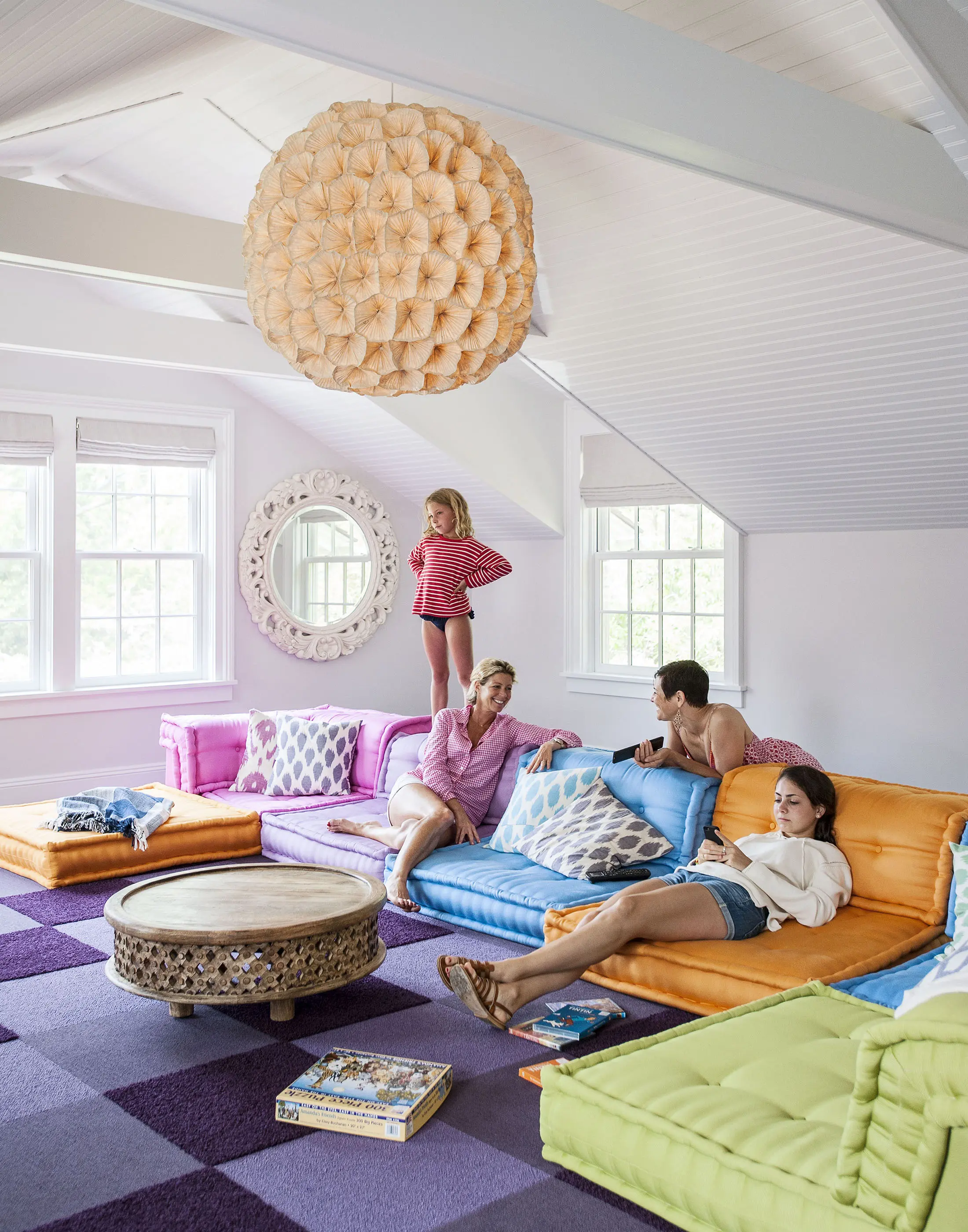 A family relaxes in a bright, cozy living room with colorful seating. A woman and a man sit on blue and orange floor sofas. A young girl stands on a pink sofa, and another woman reclines on a green one. A round wooden table is at the center. A large, decorative light hangs overhead.