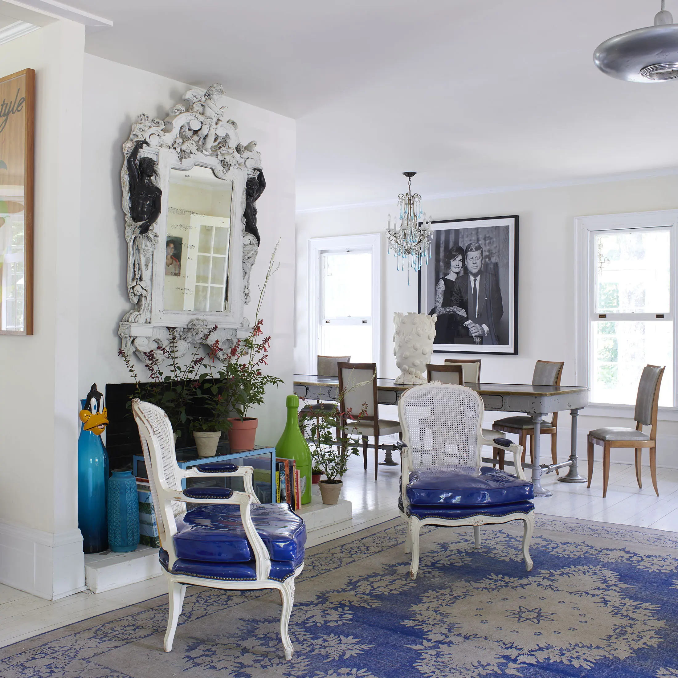 A stylish interior features two white chairs with vibrant blue cushions on a blue-patterned rug. A decorative mirror hangs on the wall near potted plants. The dining area has a table, chairs, and a chic chandelier. A large black-and-white photograph is displayed on the wall.