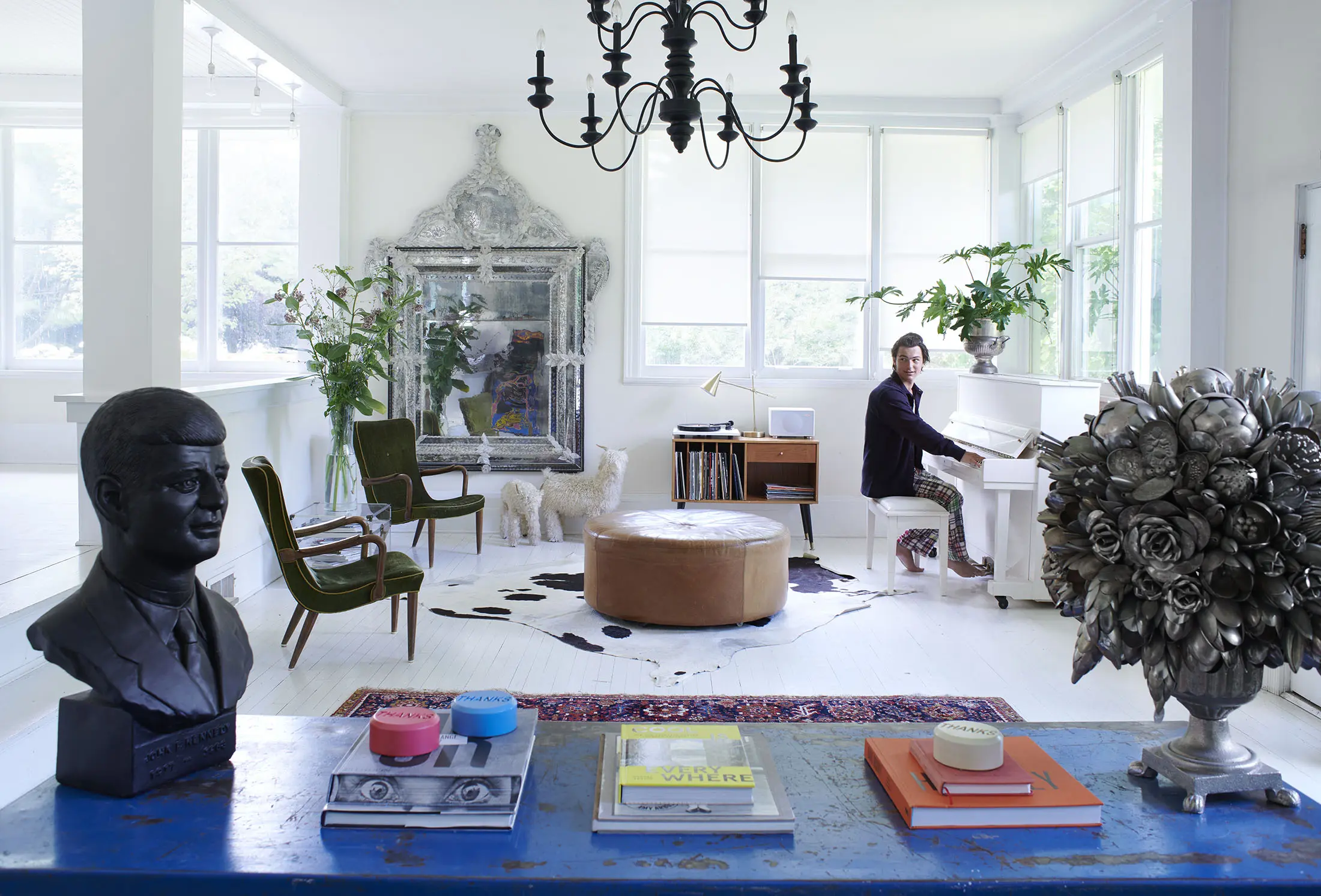 A bright, airy room with a person playing a white piano. A bust sculpture sits on a blue table in the foreground, alongside books and colorful bowls. The room has green chairs, a large mirror, potted plants, and a cowhide rug. Natural light floods through many windows under a dark chandelier.