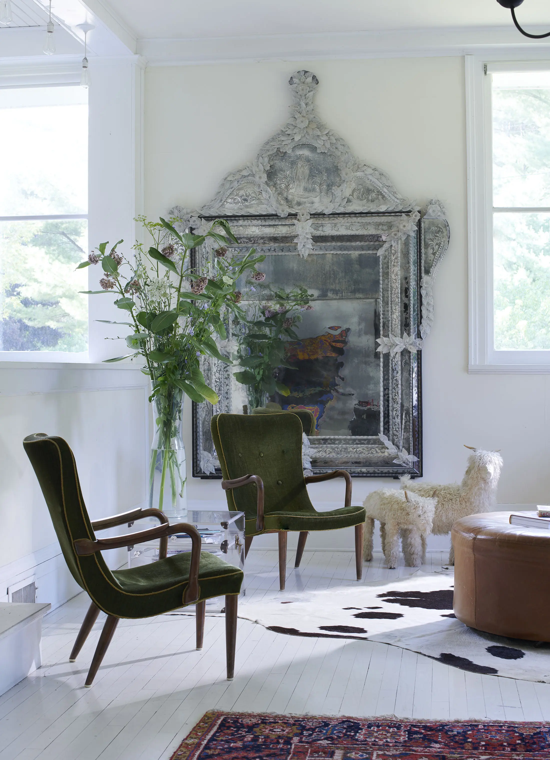 A serene room features two vintage green armchairs on a white wooden floor. Behind them, a large ornate silver mirror leans against the wall. A tall vase of flowers stands nearby. A small plush alpaca decoration is beside a brown leather ottoman, with a cowhide rug partially visible.