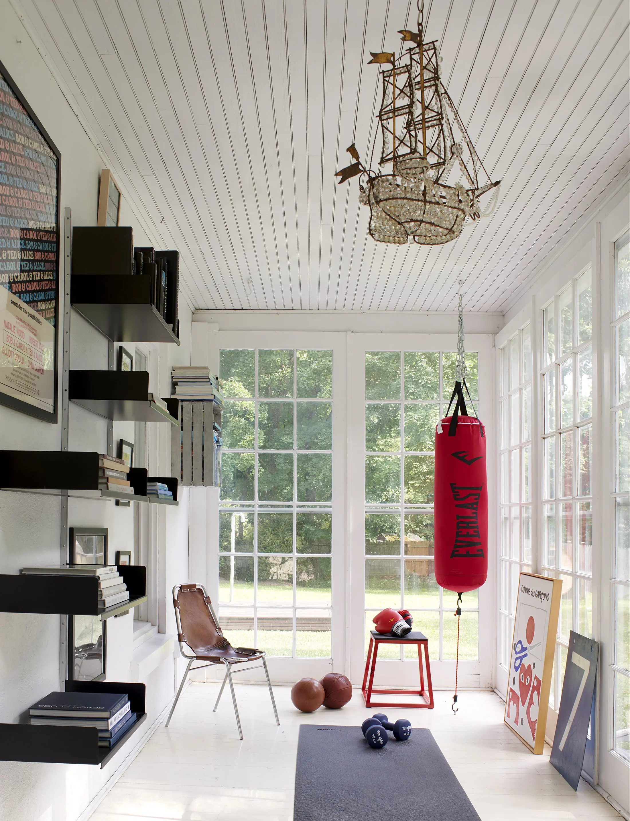 A bright room with white wooden walls and ceiling, featuring a ship-shaped chandelier. Theres a wall-mounted black shelf with books. A red punching bag, exercise equipment, and framed art are on the floor near large windows that offer a view of greenery outside.