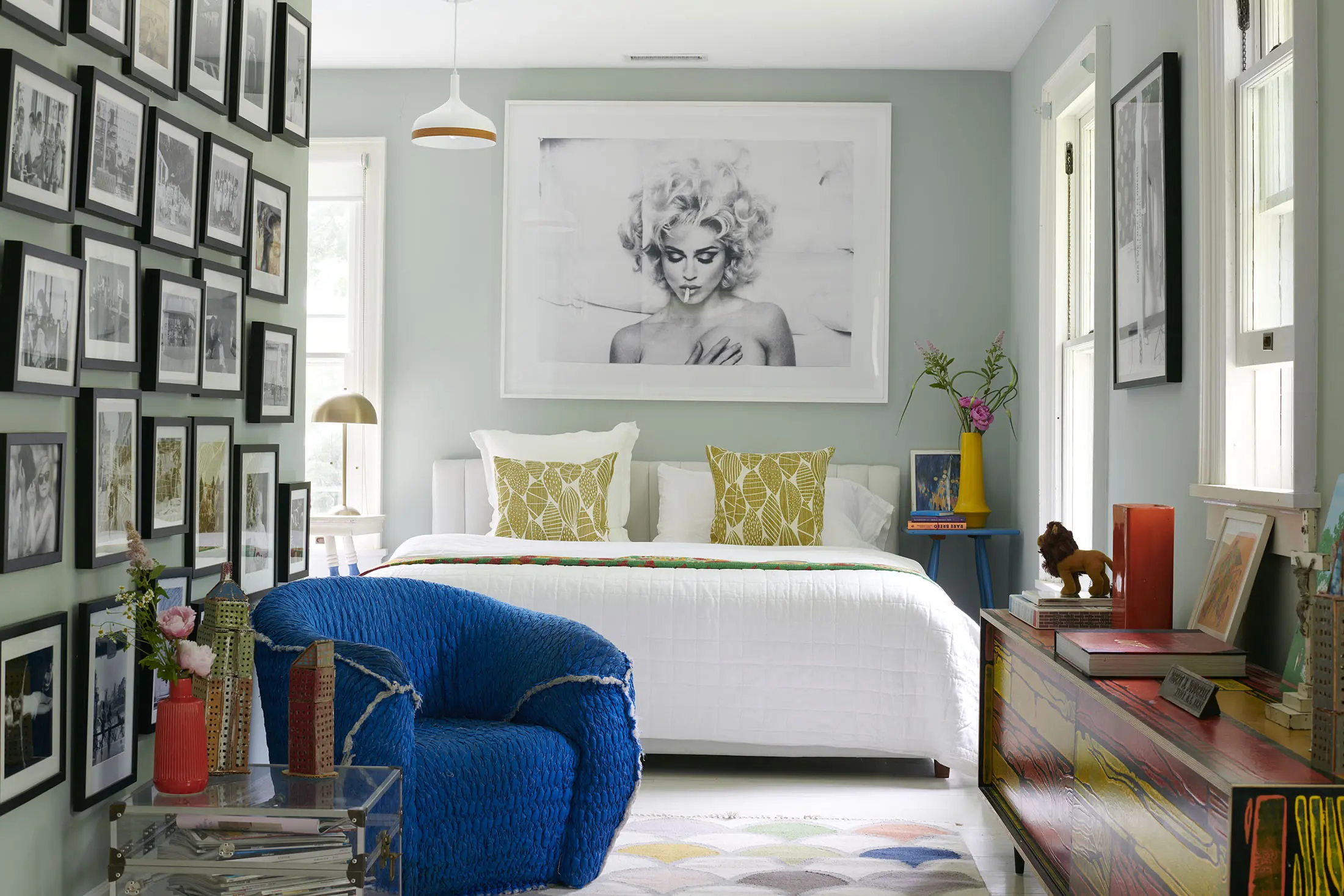 A cozy bedroom with a white bed featuring yellow-patterned pillows, under a large framed black-and-white portrait. A blue textured chair sits nearby. The room has a gallery wall of photos, colorful dresser with books, and a vase with flowers. A geometric rug covers the floor.