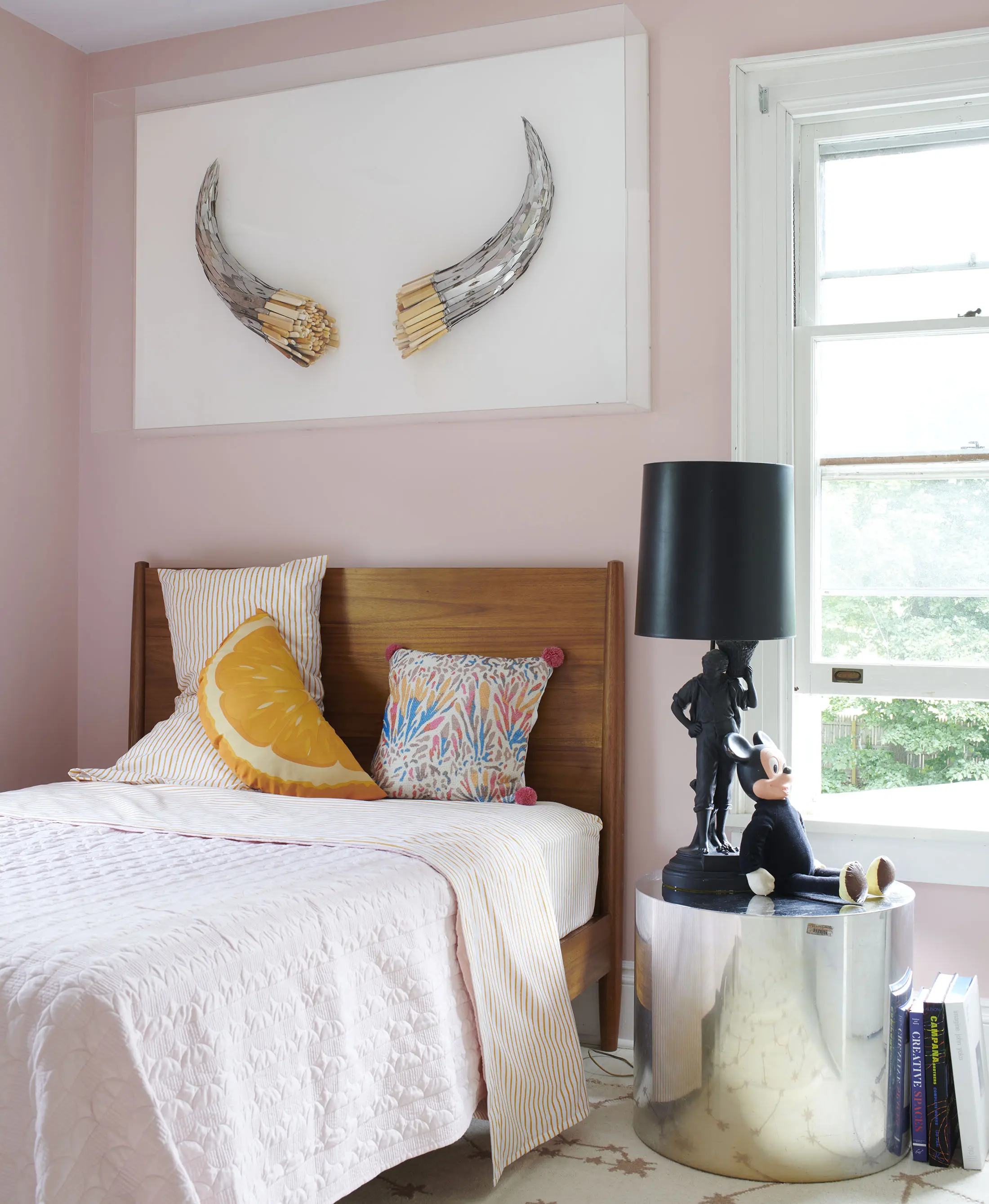 A bedroom with pale pink walls features a wooden bed with pink bedding and pillows, including yellow and patterned designs. Above the bed are decorative tusks. A silver cylindrical side table holds a black lamp and a small figurine. Books rest on the floor by the window.