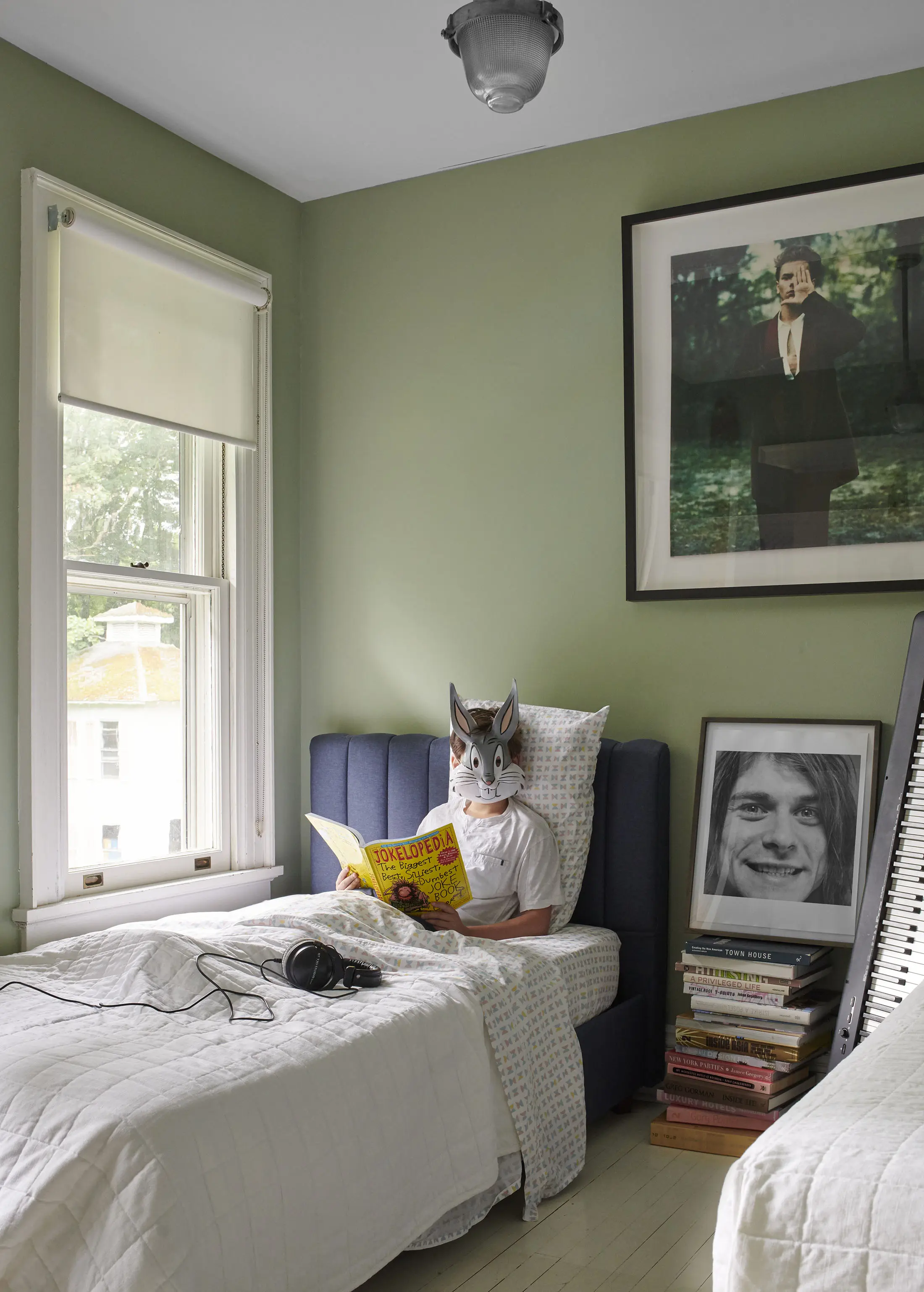 A person in a wolf mask sits on a bed reading a comic. The room has light green walls and a large framed photograph above the bed. A stack of books with a photograph of a smiling face leans against the bed. A window with a white blind is partially open, letting in natural light.