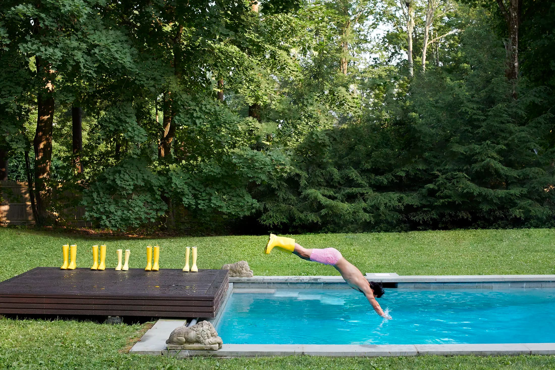 A person in pink shorts dives into a backyard pool surrounded by trees. Several yellow rain boots are lined up on a wooden deck beside the pool. The scene is sunny, and the grass is green and well-kept.