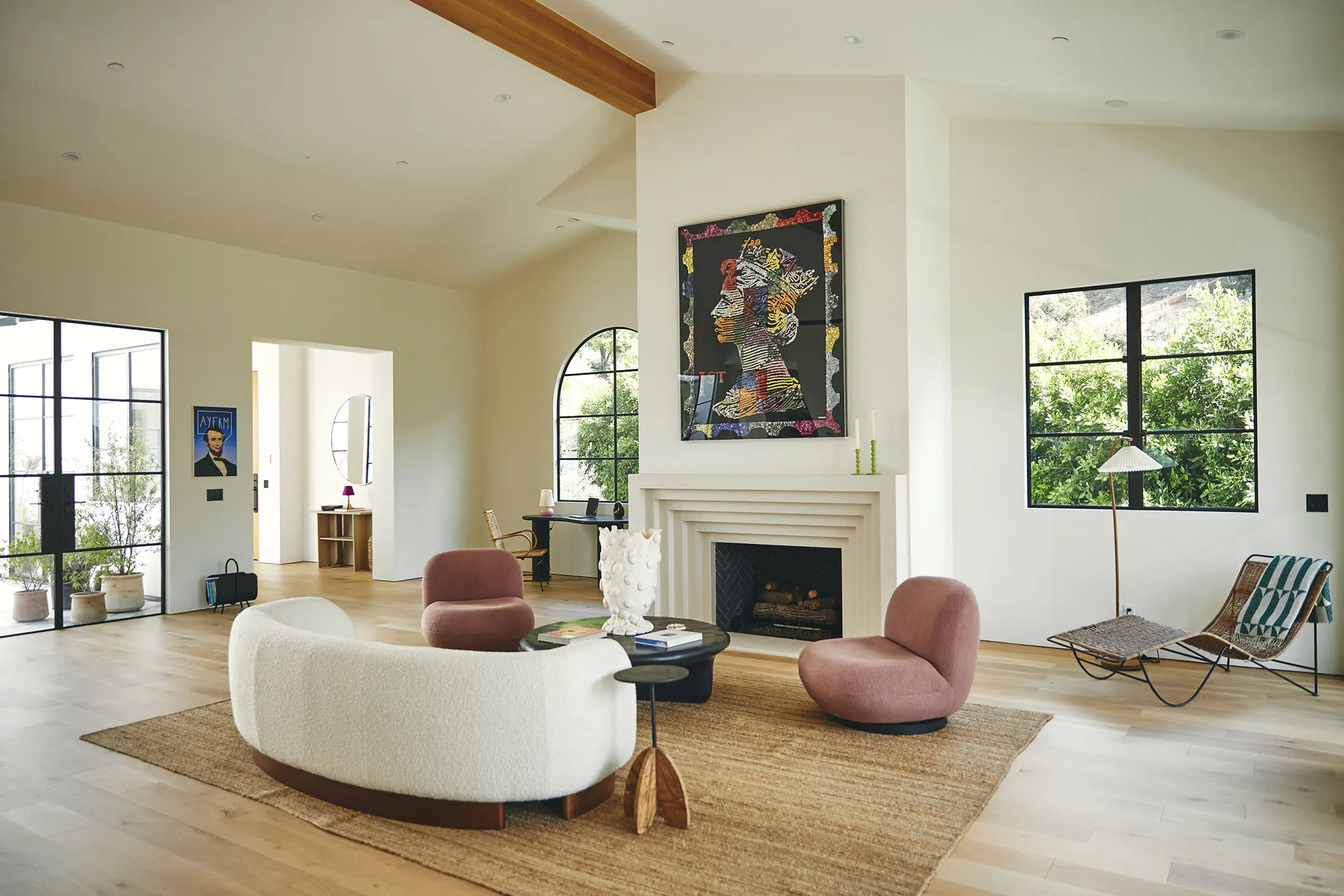 A modern, open courtyard with large glass doors leading outside. The area features a mix of wooden and metal furniture, including a table with chairs. Various potted plants are placed throughout the space. The interior has a neutral color palette with light wood flooring and white walls.