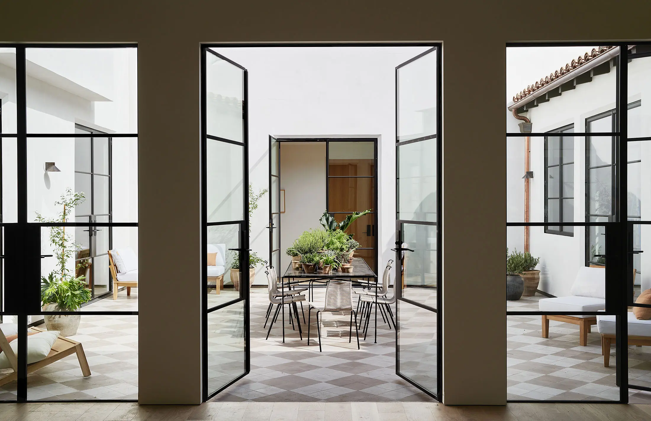 Aerial view of a courtyard with a checkered tile floor in shades of brown and beige. The courtyard is bordered by walls with multiple windows and topped with terracotta roof tiles. It features a central dining table, chairs, potted plants, and seating in the corners.