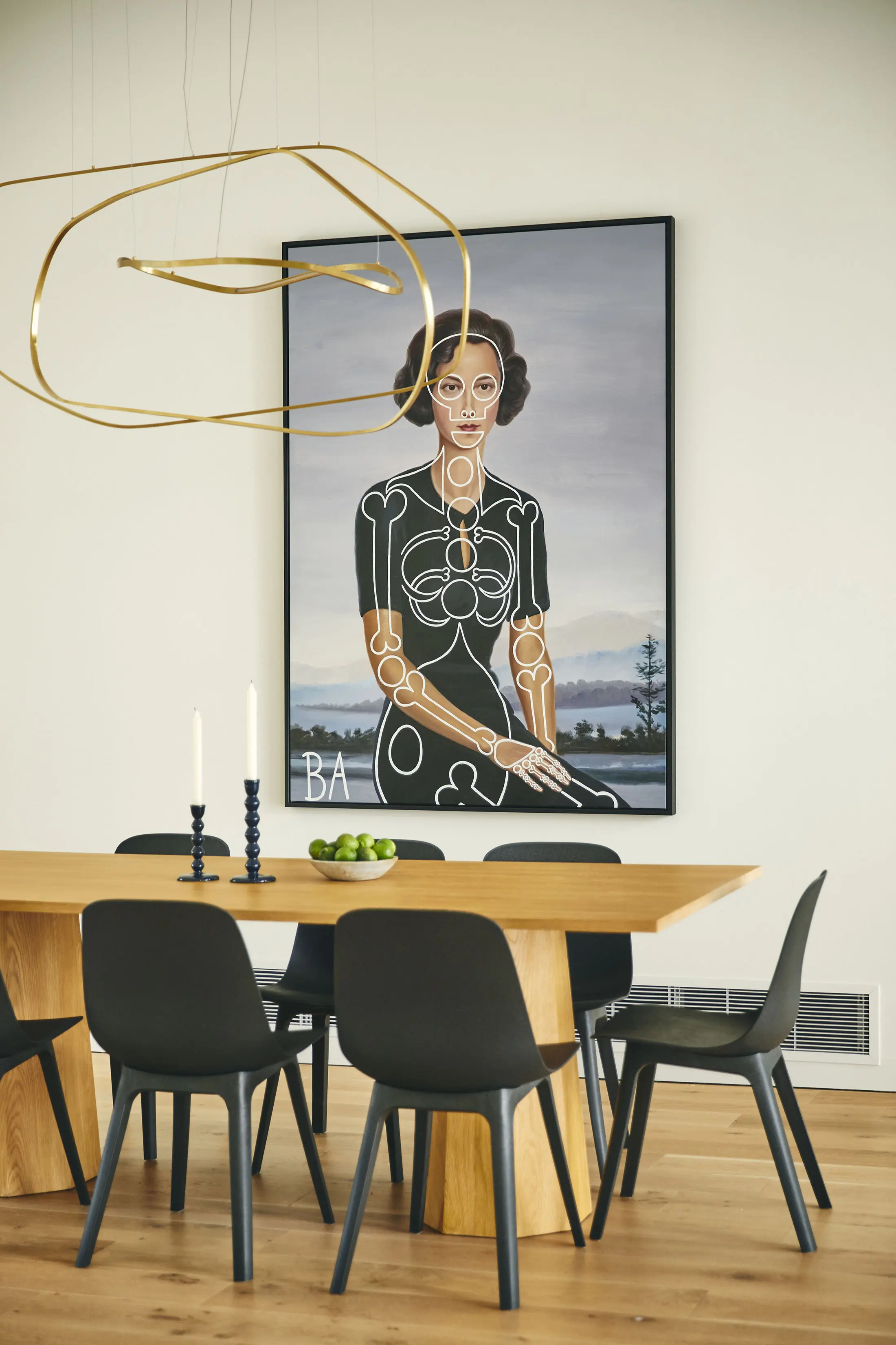 A modern dining room features a wooden table with six black chairs. On the table are two white candles and a bowl of green apples. Above hangs a geometric gold light fixture. A large, stylized portrait of a woman in a unique outfit is on the wall in the background.