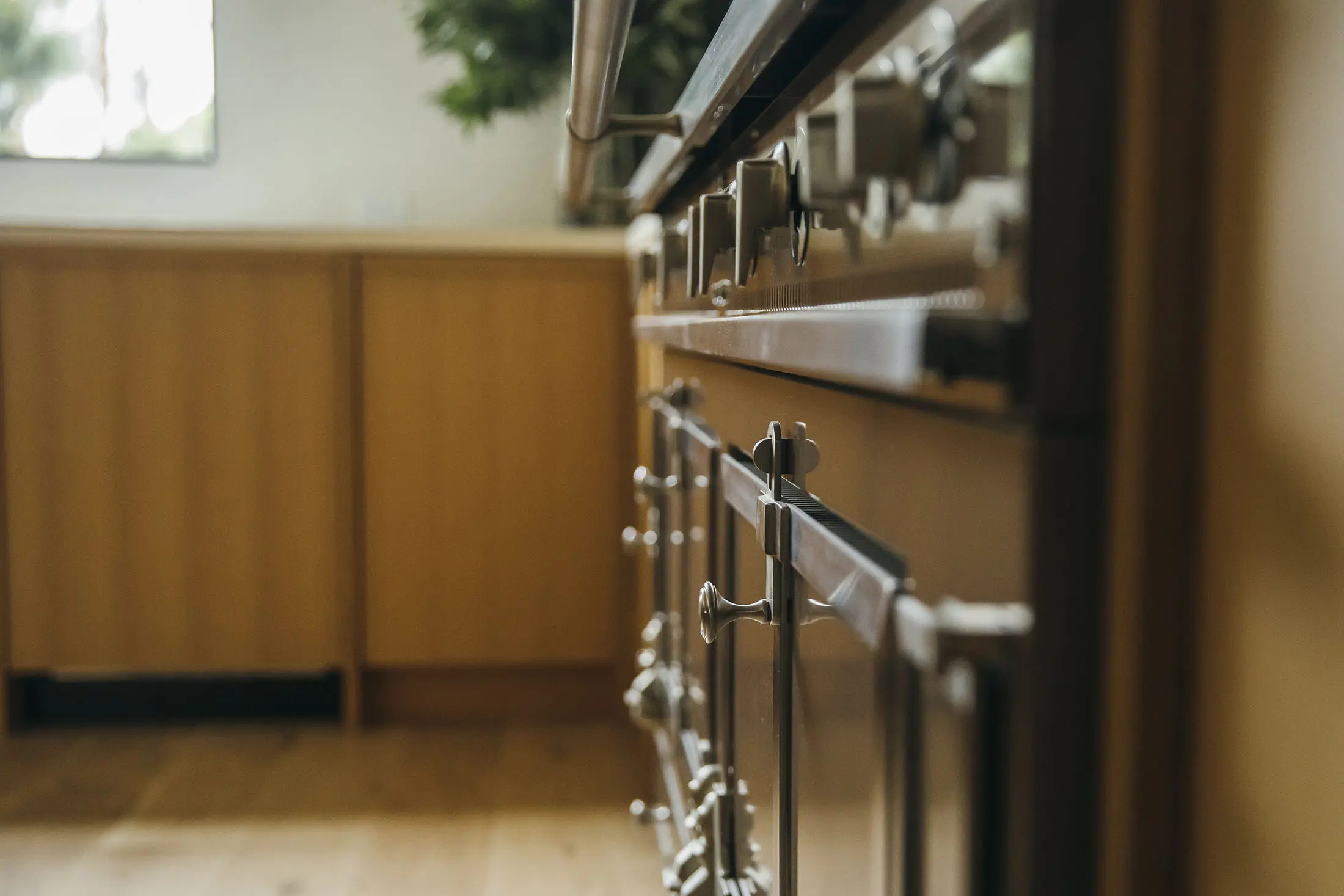 Close-up of a kitchen with a focus on the metallic knobs of a stove on the right. The cabinetry is wooden, with a blurry background showing more kitchen cabinets and a small window. The floor is wooden, adding warmth to the modern kitchen setting.