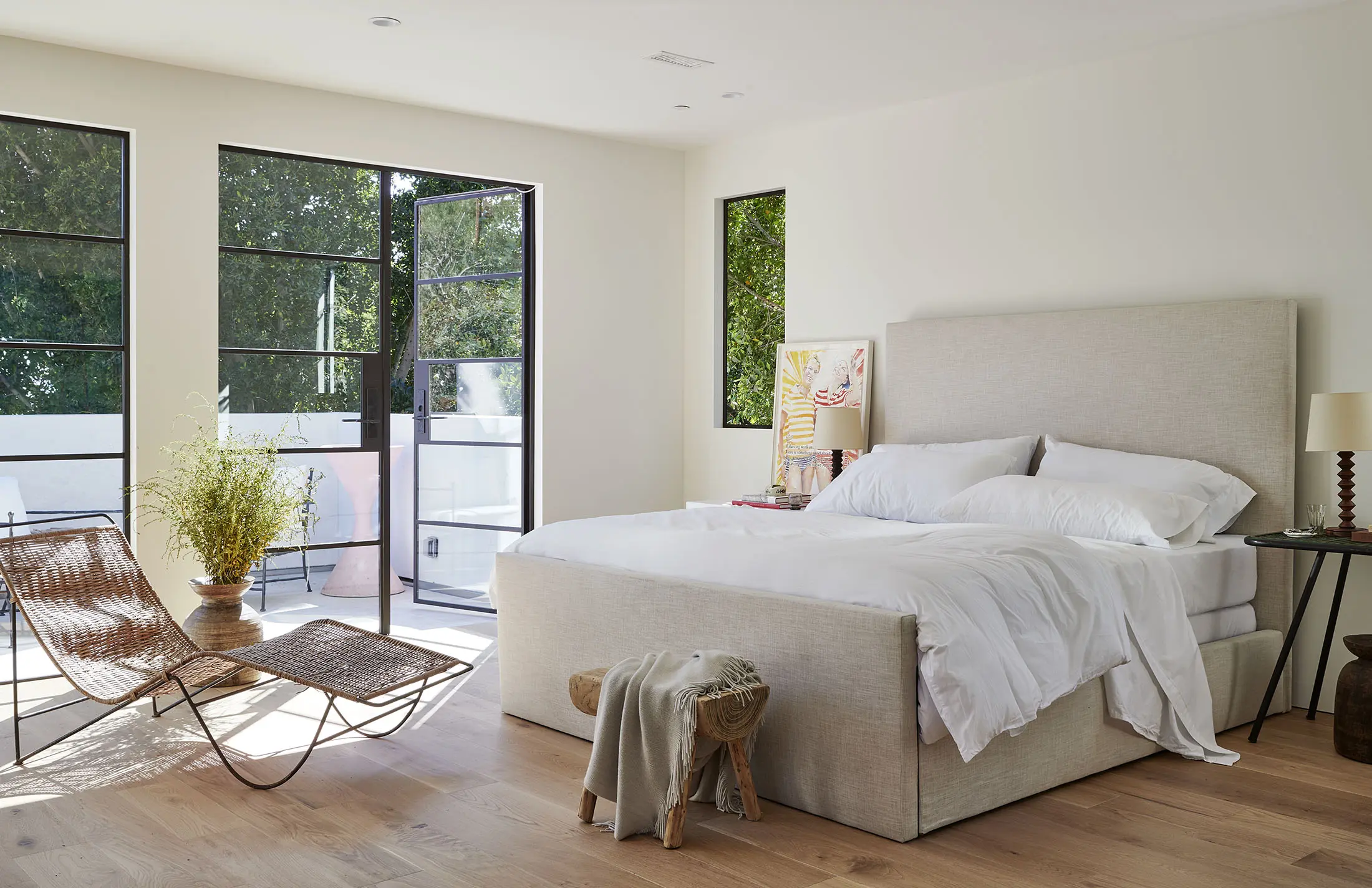 A bright bedroom with a large upholstered bed and white bedding. To the left, two woven chairs face a glass door opening to a balcony with greenery. A wooden stool with a draped blanket sits near the bed. A side table with a lamp and books is by the window. Light wood flooring.