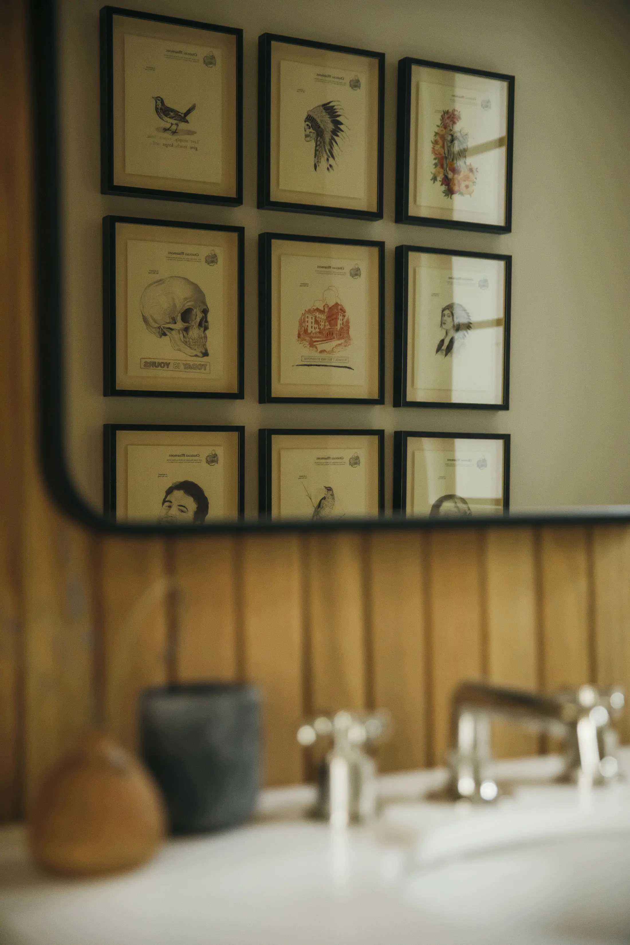 A bathroom mirror reflects a gallery wall with nine framed artworks. The images include a bird, an eagle feather headdress, a floral design, a human skull, and various artistic sketches. In the foreground, a sink with a silver faucet and two containers on the counter are visible.