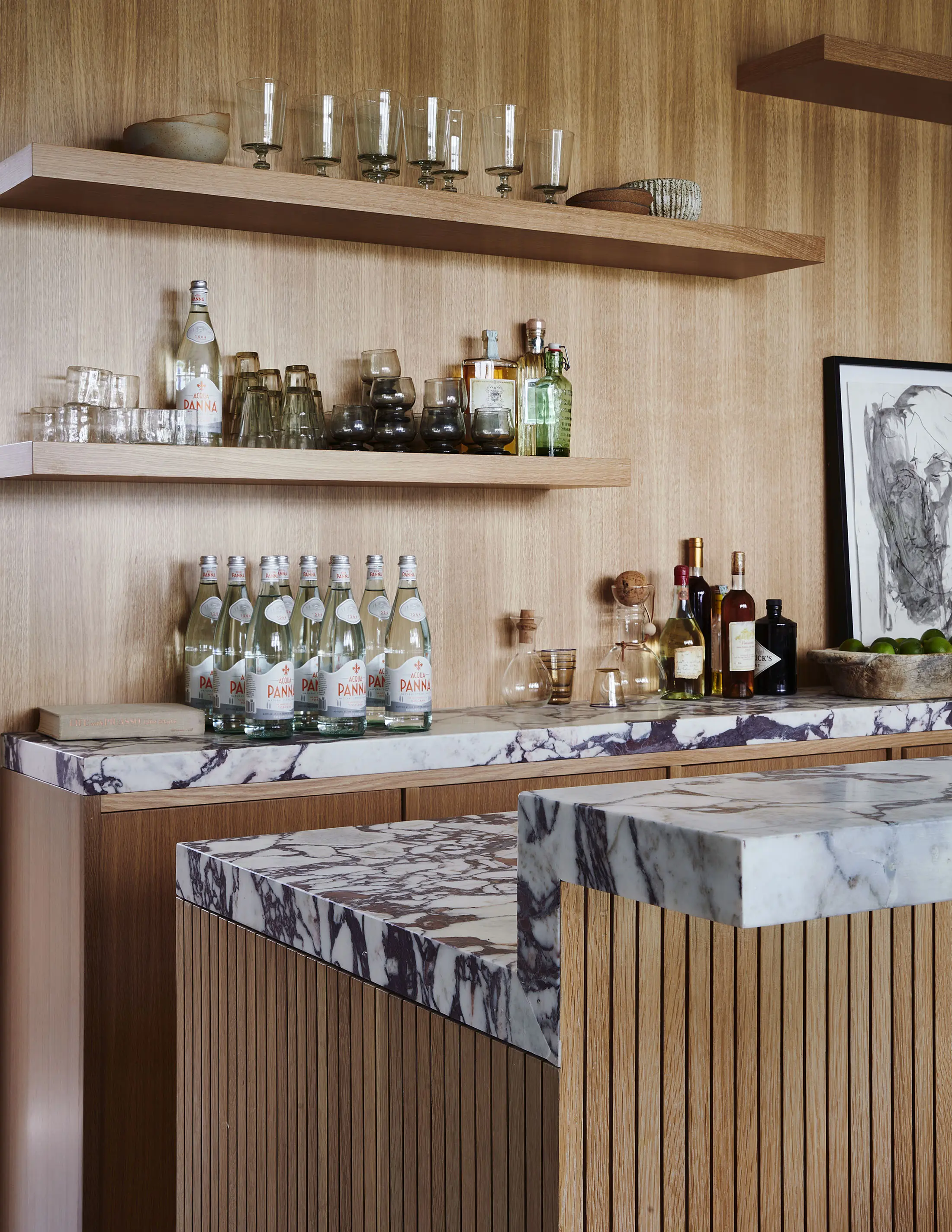 Modern kitchen bar with wooden paneling and marble countertops. Open shelving displays glasses, mineral water bottles, liquor bottles, and decorative items. A framed black-and-white abstract artwork leans on the counter, alongside a bowl of limes and a bottle of olive oil.