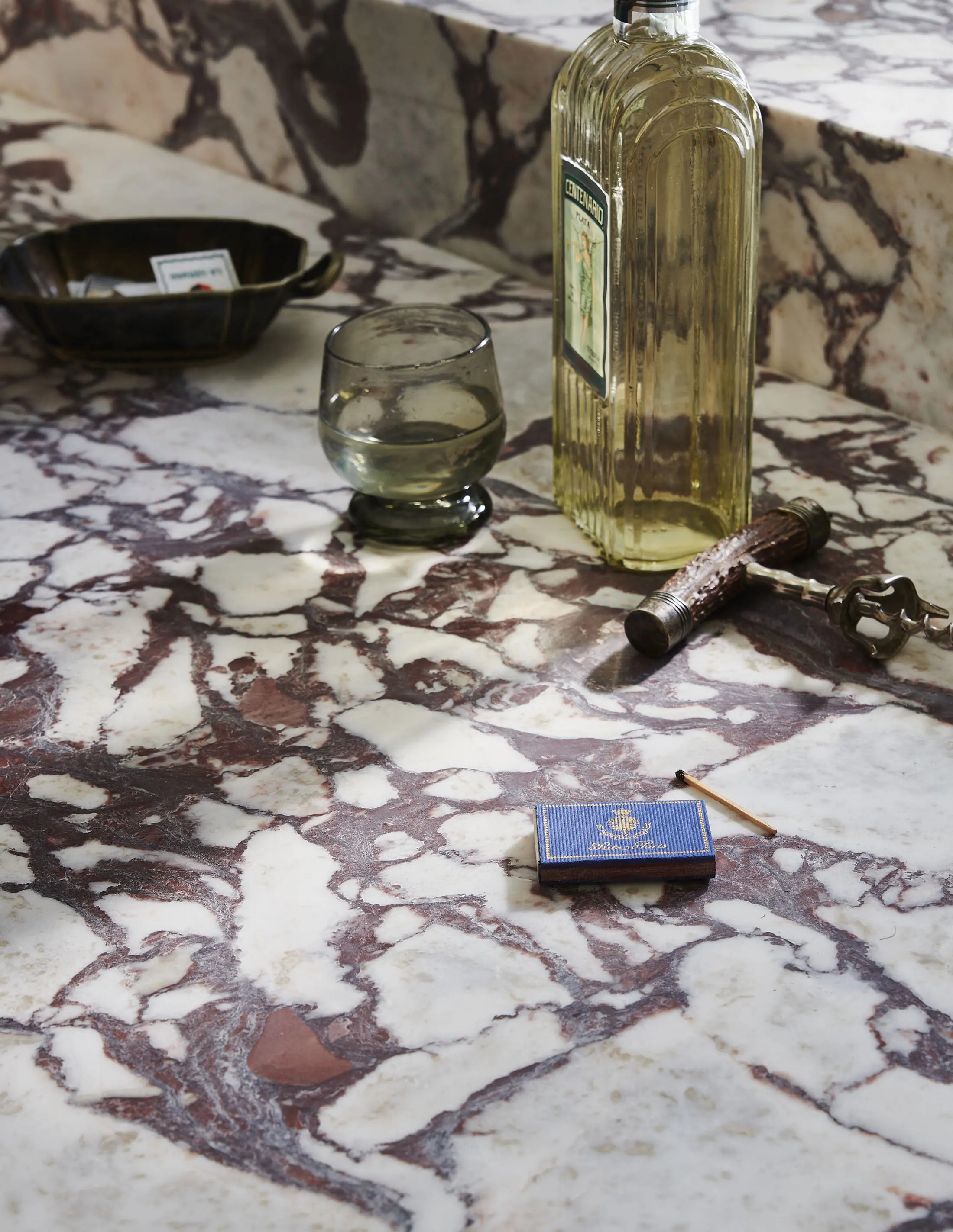 A mottled marble surface with brown, gray, and white patterns holds a green glass cup, an open matchbox, a vintage key, and a vintage bottle of clear liquid. The scene suggests a rustic, vintage aesthetic with soft lighting highlighting the intricate details of the marble.