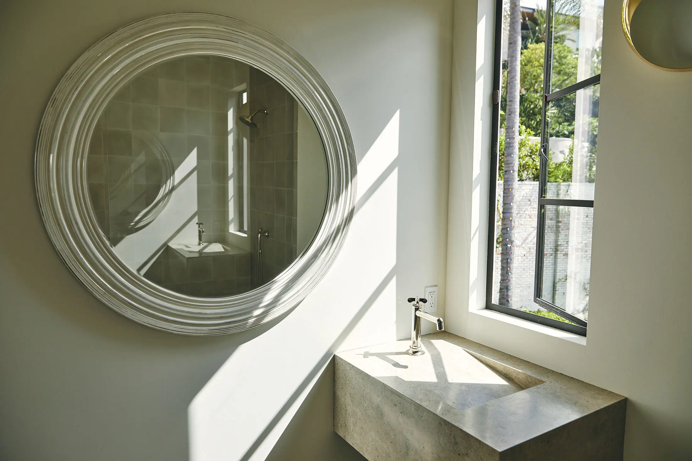 A minimalist bathroom features a circular mirror above a sleek, rectangular stone sink with a stainless steel faucet. Sunlight streams through a large window on the right, casting shadows on the pale wall. Outside, green foliage and trees are visible.