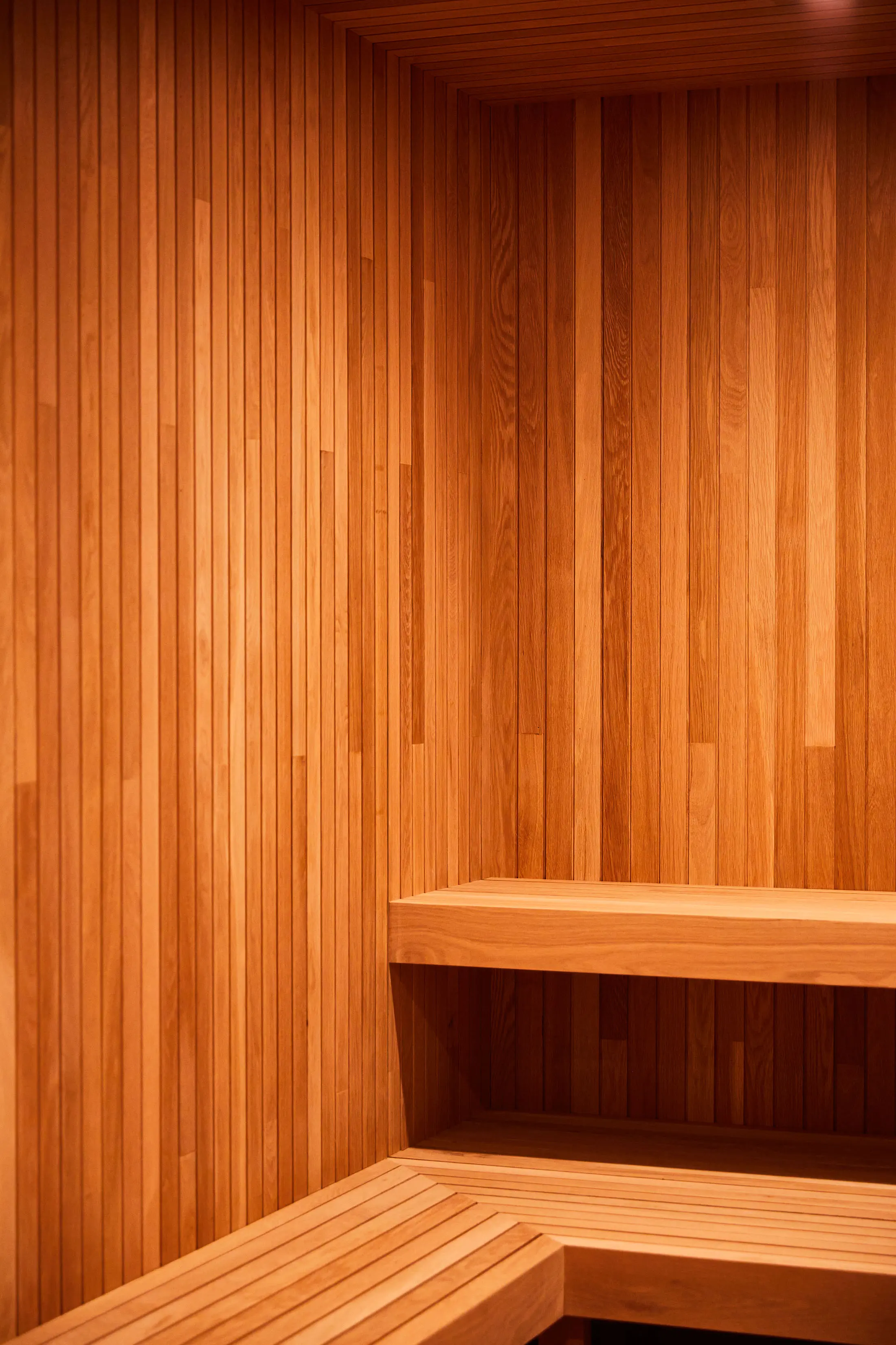 Wooden sauna interior with vertical slatted walls and two-tiered benches. The warm, honey-colored wood creates a cozy atmosphere. Light from above highlights the natural grain and texture of the wood, evoking a sense of calm and relaxation.