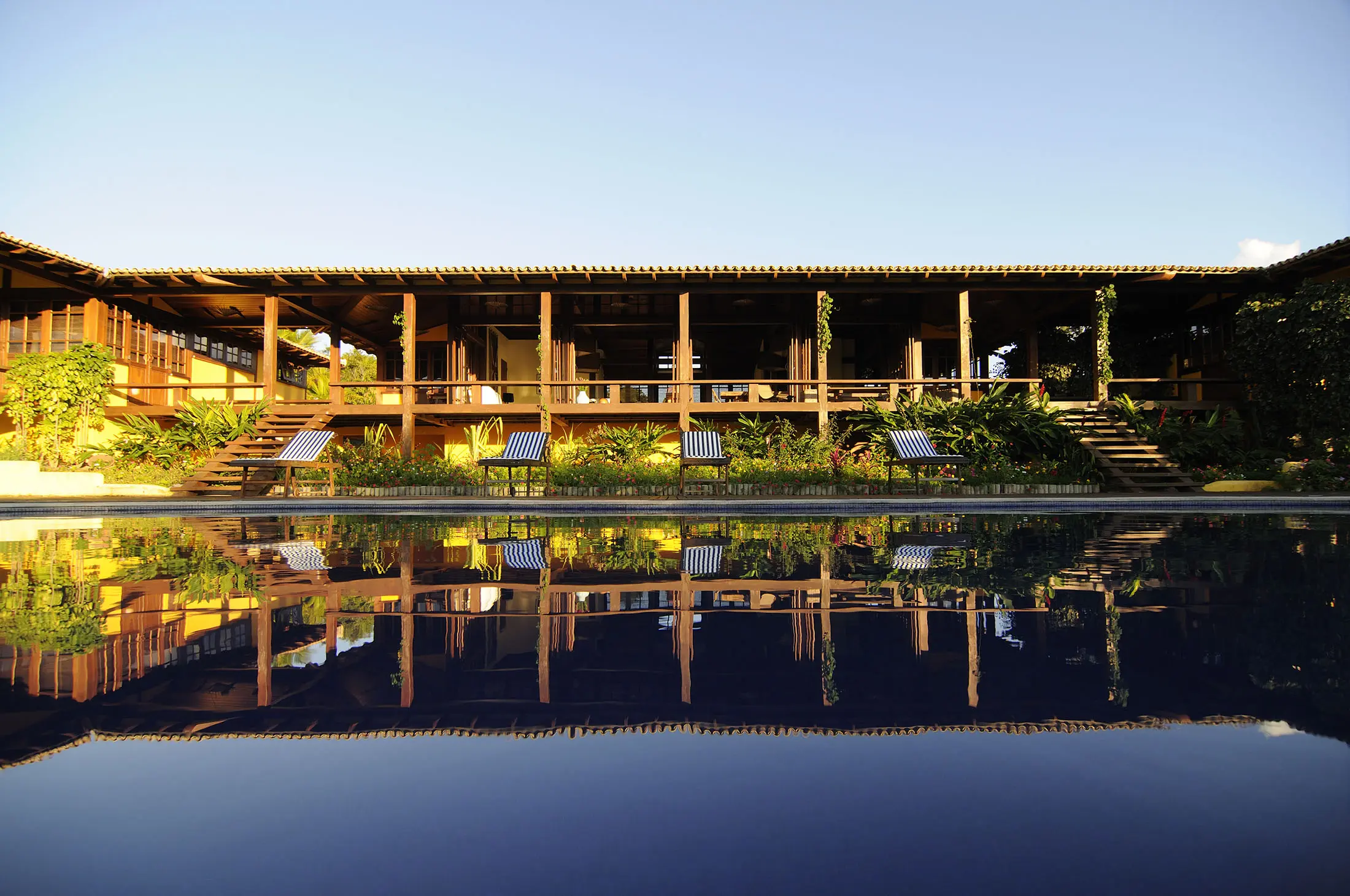 A large wooden house with a thatched roof, featuring open sides and surrounded by greenery, reflects on a calm swimming pool in the foreground. Wooden lounge chairs with striped cushions are lined up near the pool. The sky is clear and blue, suggesting a sunny day.