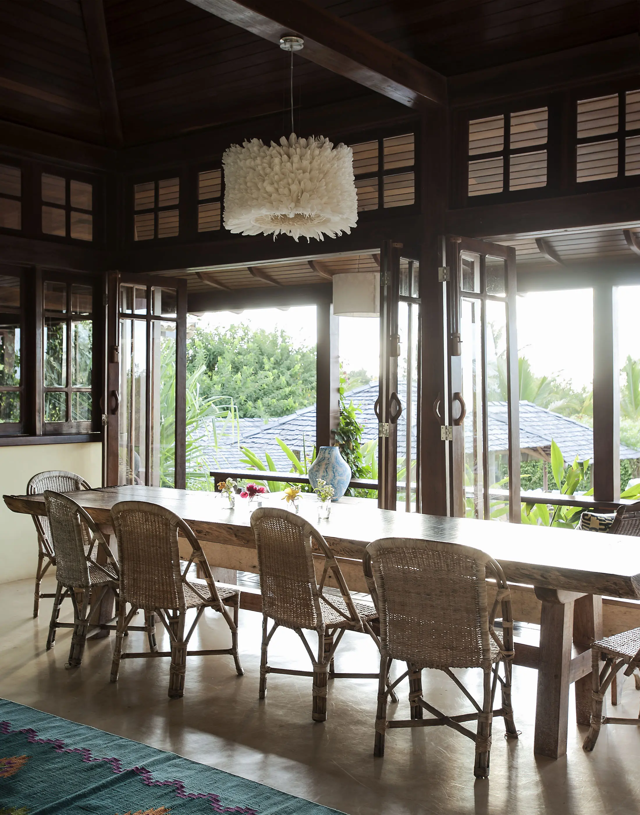Spacious dining area with wooden table and seven wicker chairs facing glass doors opening to a lush garden view. A decorative, white, feather-like chandelier hangs from the wooden ceiling. Natural light fills the room, highlighting the modern, earthy decor and green landscape outside.