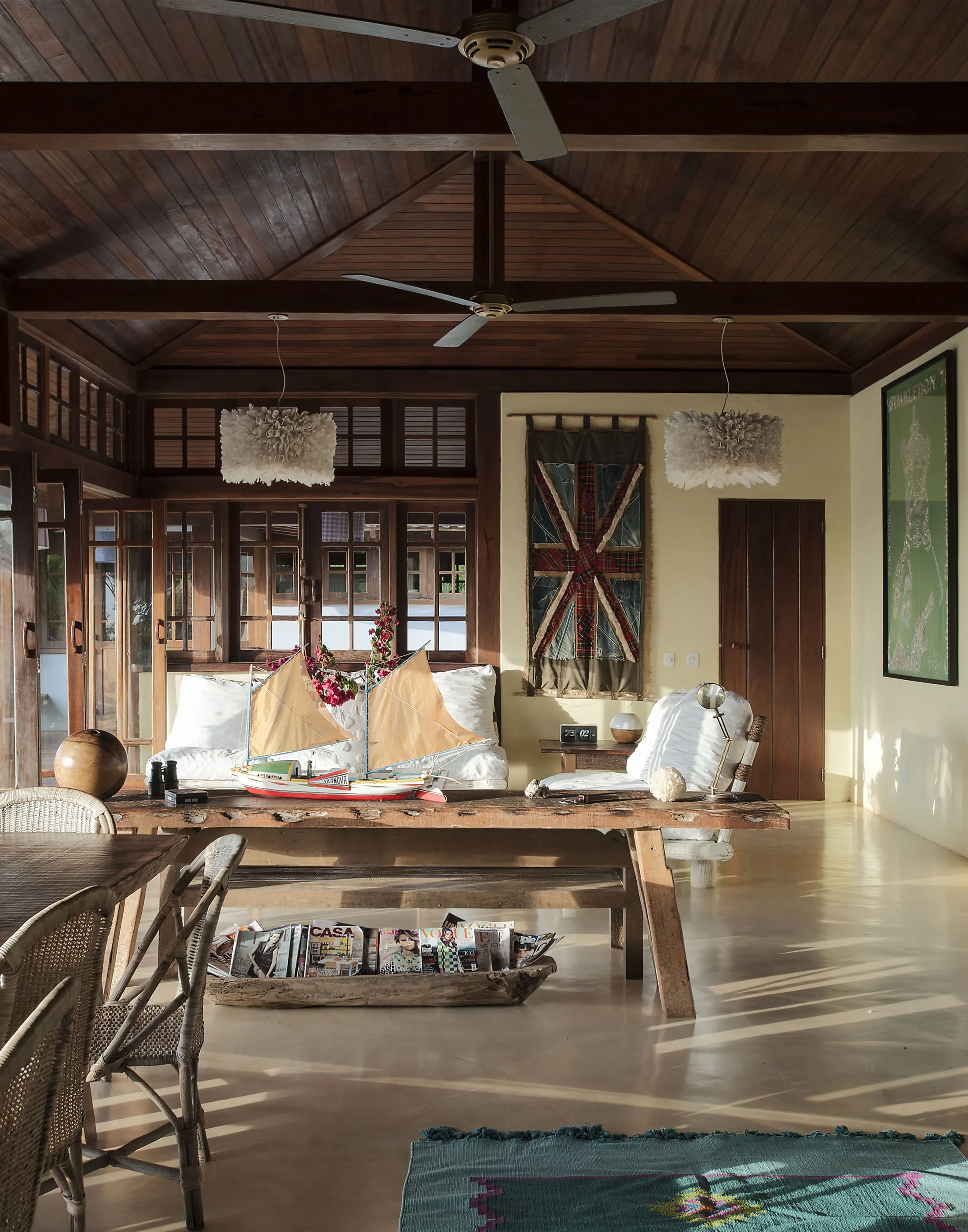 Spacious living room with wooden ceiling and beige walls. Features a long wooden table and benches, with cushions and decorative bowls. Two pendant lights hang, resembling fans. A British flag fabric decorates one wall. Sunlight streams in through large windows, illuminating the space.