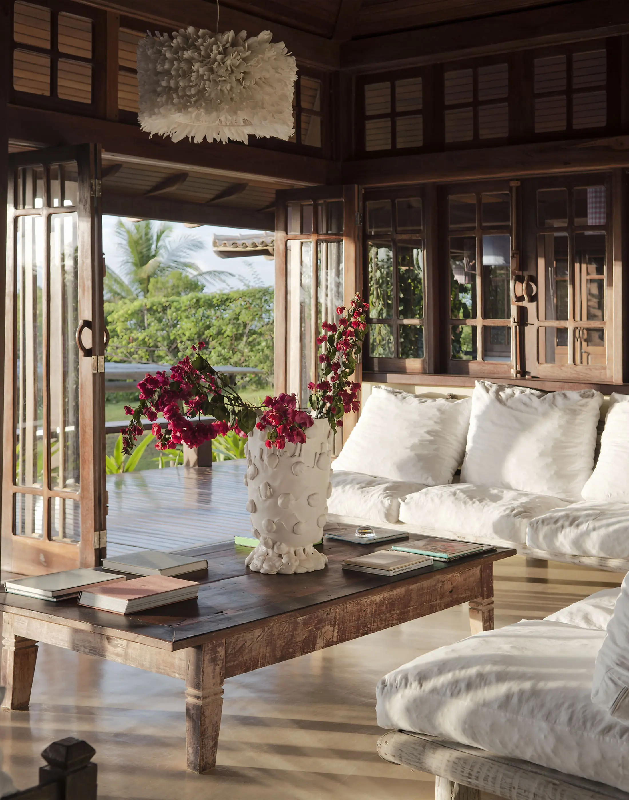 A cozy living room with wooden paneling features an open doorway leading to a lush garden. A rustic wooden table holds a white vase filled with vibrant pink flowers, flanked by books. Plush white cushions adorn the surrounding sofas. A decorative white pendant light hangs above.