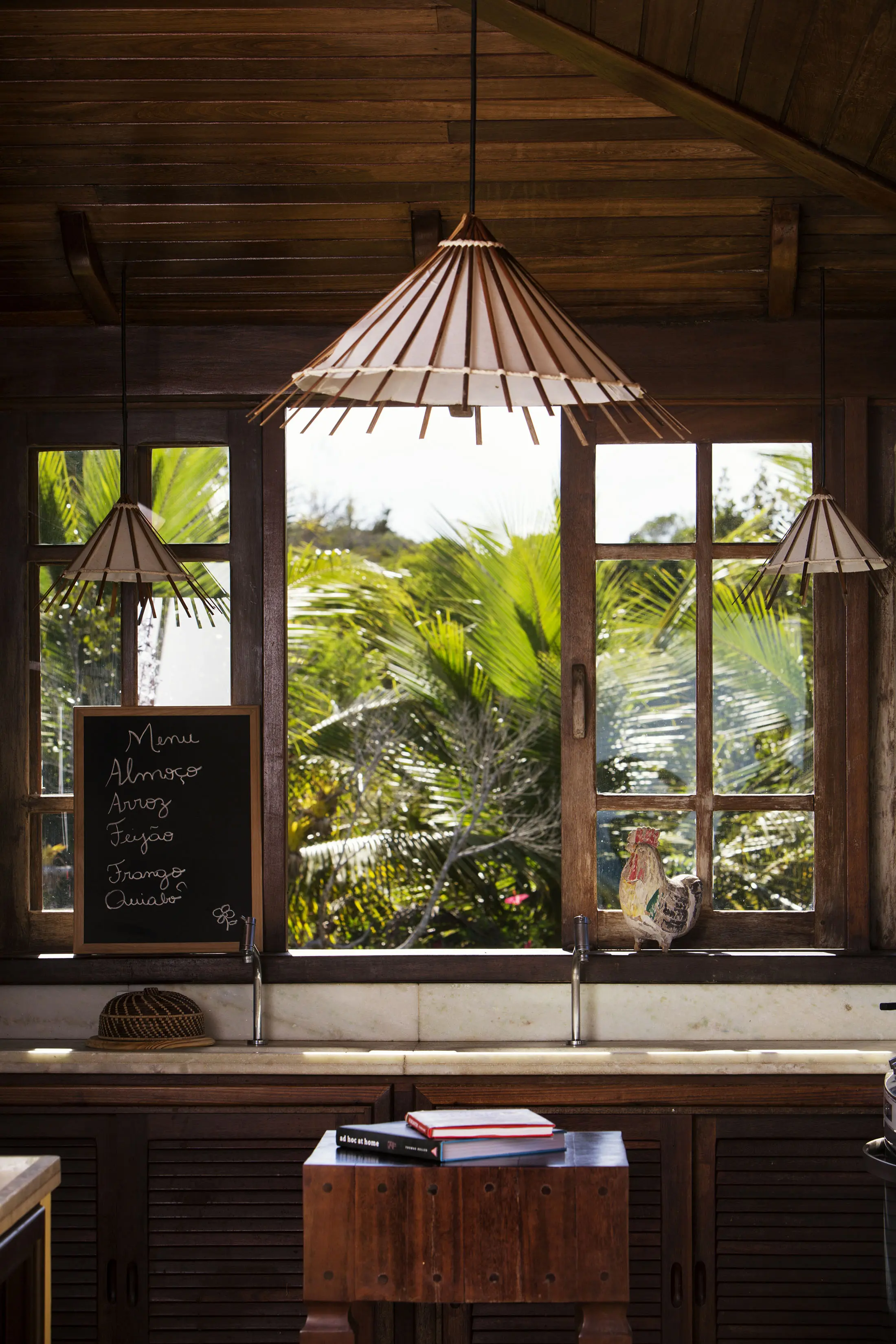 Rustic kitchen interior with wooden beams and large windows. Three pendant lamps with conical shades hang from the ceiling. A chalkboard with a Spanish menu stands by the sink. Outside, lush green palm trees are visible. A ceramic rooster decorates the windowsill.