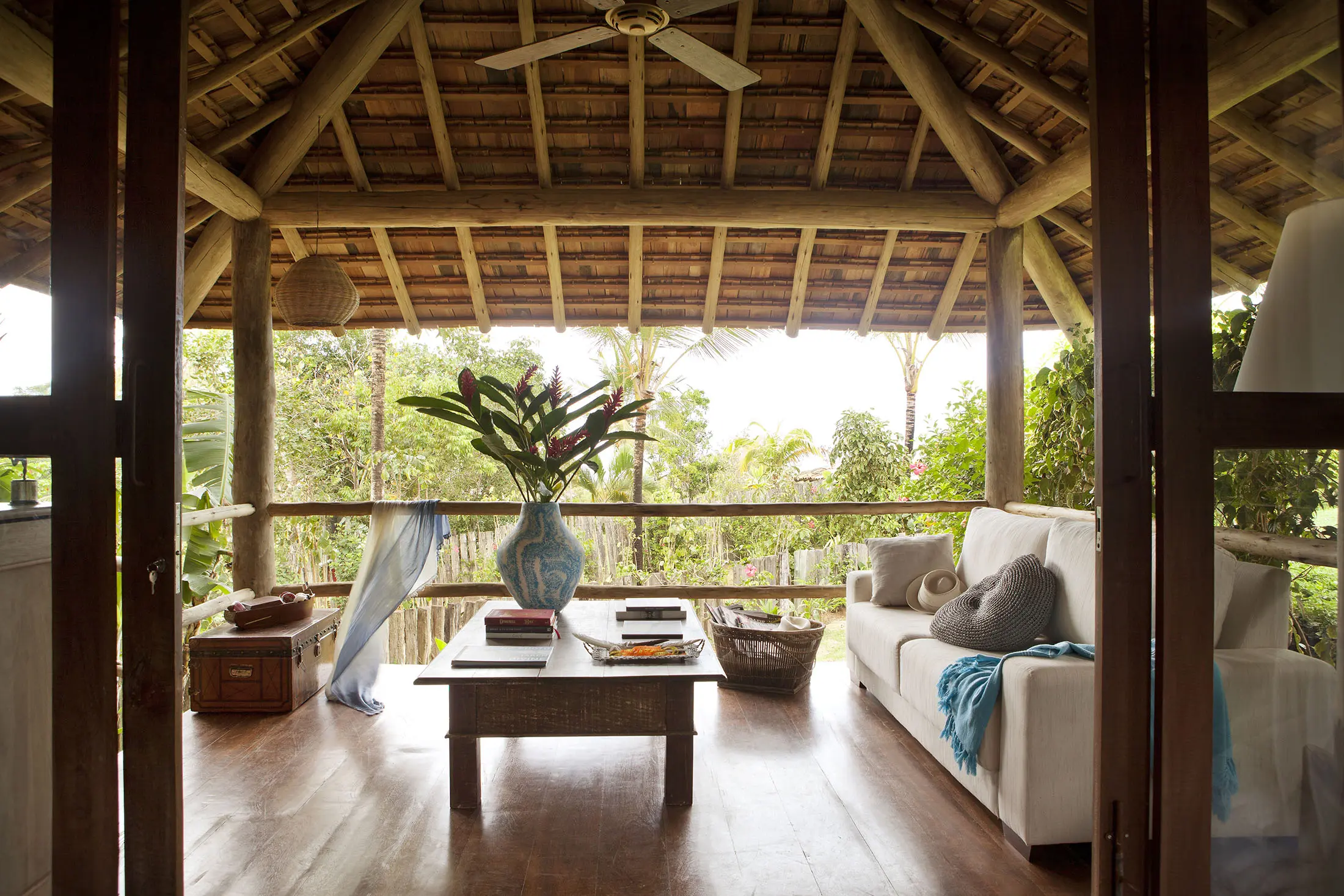 Rustic outdoor living space with a wooden ceiling and floor, featuring a white sofa adorned with blue blankets and pillows. A wooden coffee table displays a large vase with tropical leaves. Sheer blue curtains gently drape from the ceiling, and lush greenery surrounds the area.