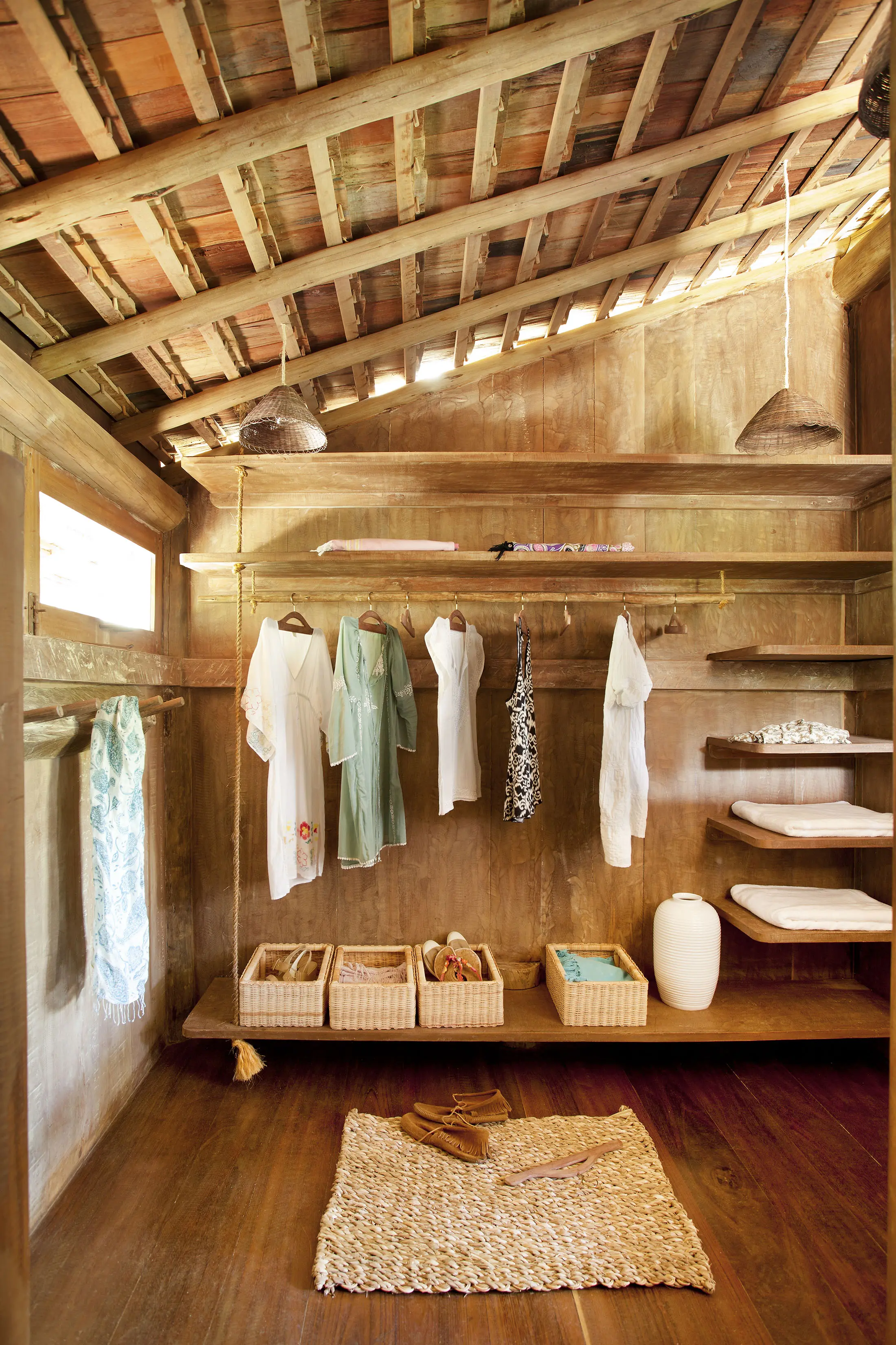 Rustic wooden closet with open shelving and a slanted roof. Clothes, including dresses and shirts, hang on a rod. Baskets on the shelves store items. A woven rug lies on the wooden floor with sandals. Natural light filters through a small window, creating a warm atmosphere.