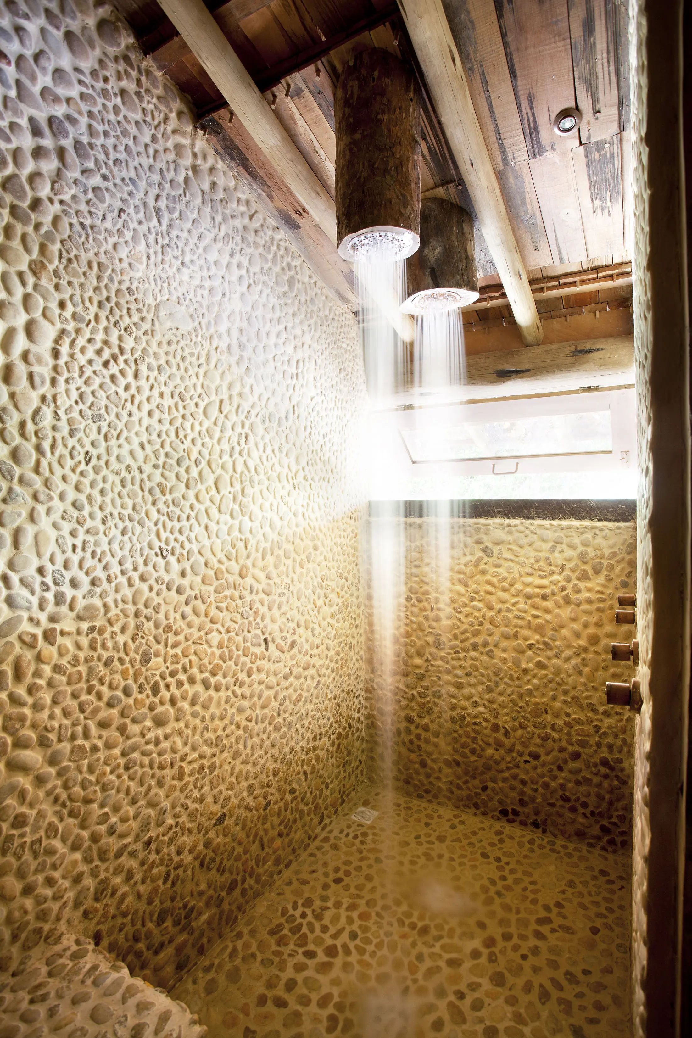 Rustic shower with textured pebble walls and a wooden ceiling. Water falls like a gentle cascade from two cylindrical wooden showerheads, creating a natural ambiance. Sunlight streams through a small window, enhancing the earthy tones and serene atmosphere.