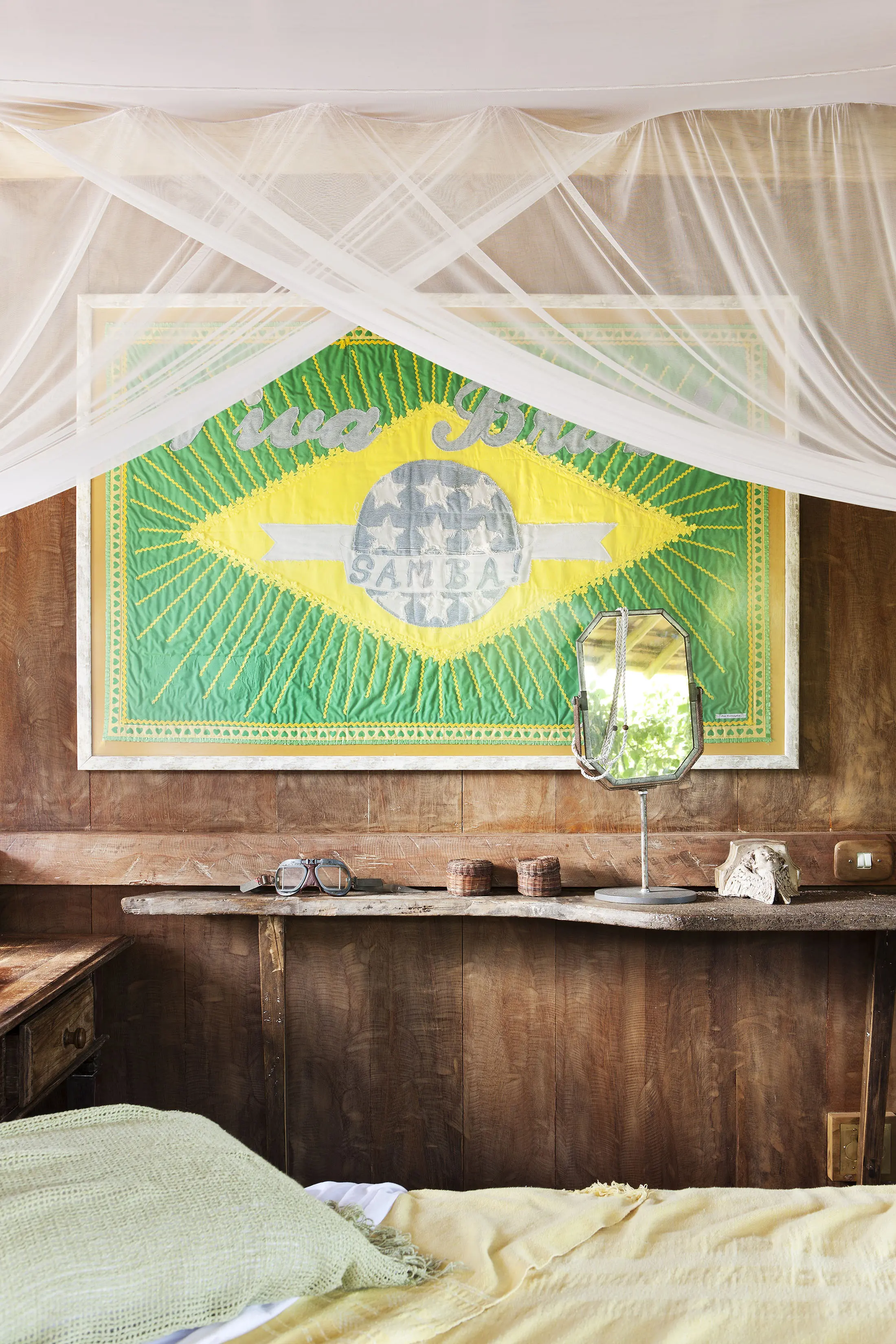 Cozy bedroom with wooden walls, featuring a large green and yellow artwork resembling the Brazilian flag. A shelf holds a mirror, two small jars, and a decorative conch shell. A soft bed with green and yellow linens is under a draped, translucent white canopy.