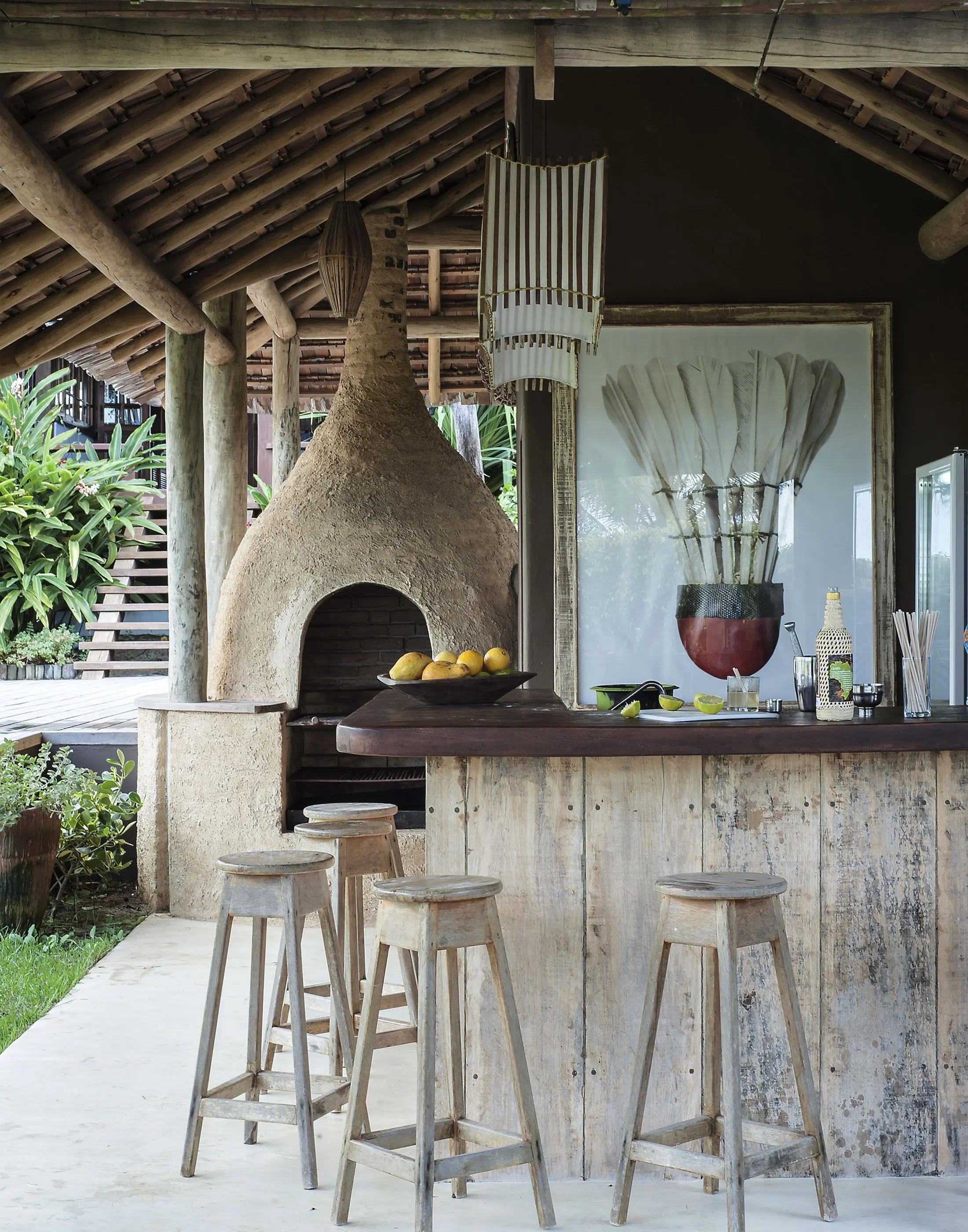 Rustic outdoor bar area with wooden stools and a countertop. Behind is a clay pizza oven with a chimney, under a slanted wooden roof. A large framed artwork of a vase with feathers hangs on the dark wall. Lemons and bottles are on the counter, with lush greenery in the background.